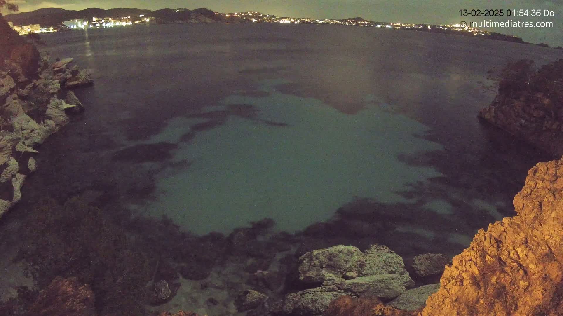 A nighttime view of a dark, calm body of water partially illuminated, surrounded by rocky cliffs and a distant, faintly lit town.