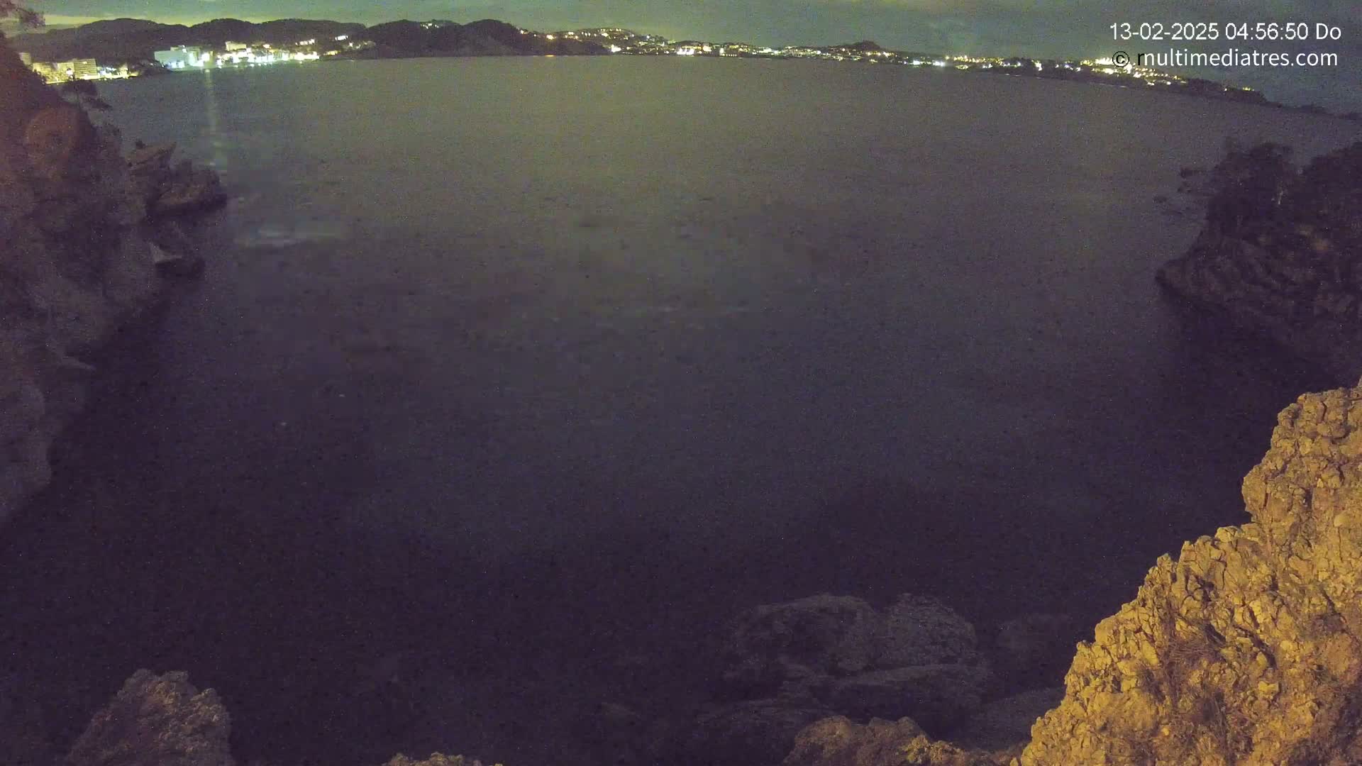 A nighttime view of a dark, calm body of water, with rocky cliffs on either side and a distant, faintly lit cityscape visible across the water.