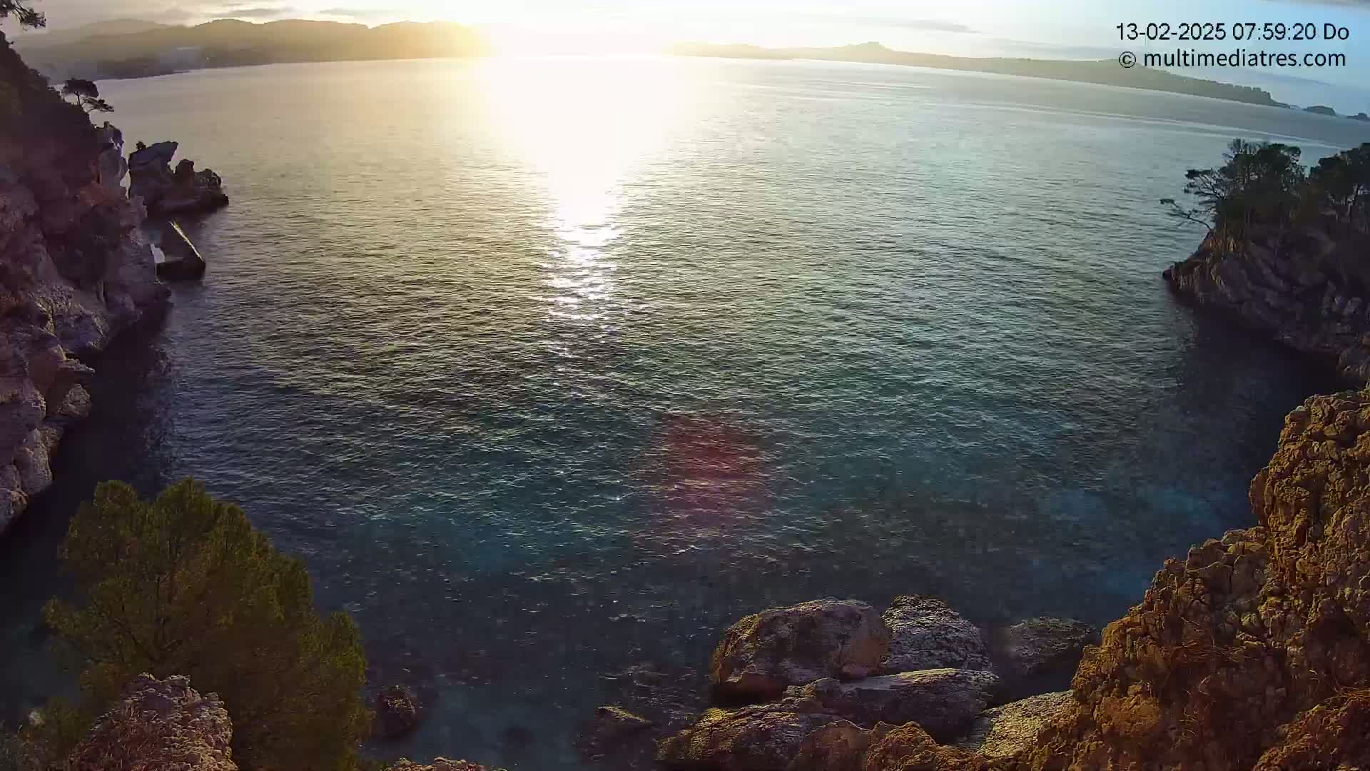 A tranquil cove at sunrise is nestled between rocky cliffs, the sun reflecting brightly on the calm, dark-blue water.