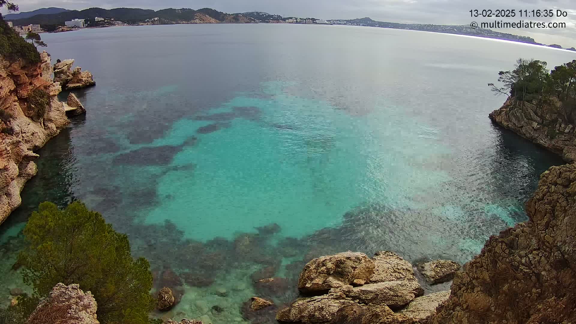 A cove of clear, teal water is nestled between rocky, tan-colored cliffs under an overcast sky.