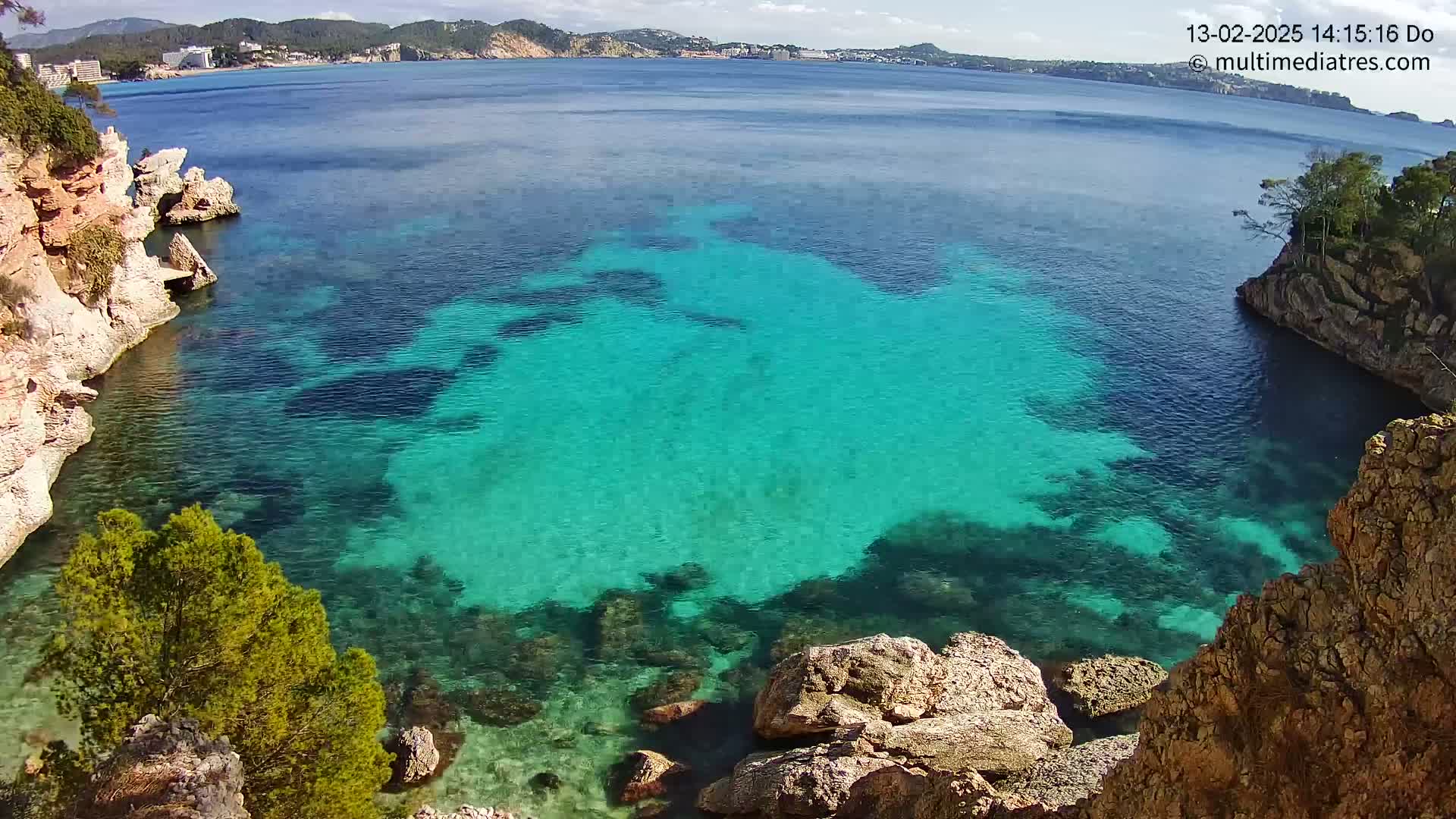 A cove of clear, turquoise water is nestled between rocky cliffs on a sunny day, with a distant coastline visible in the background.