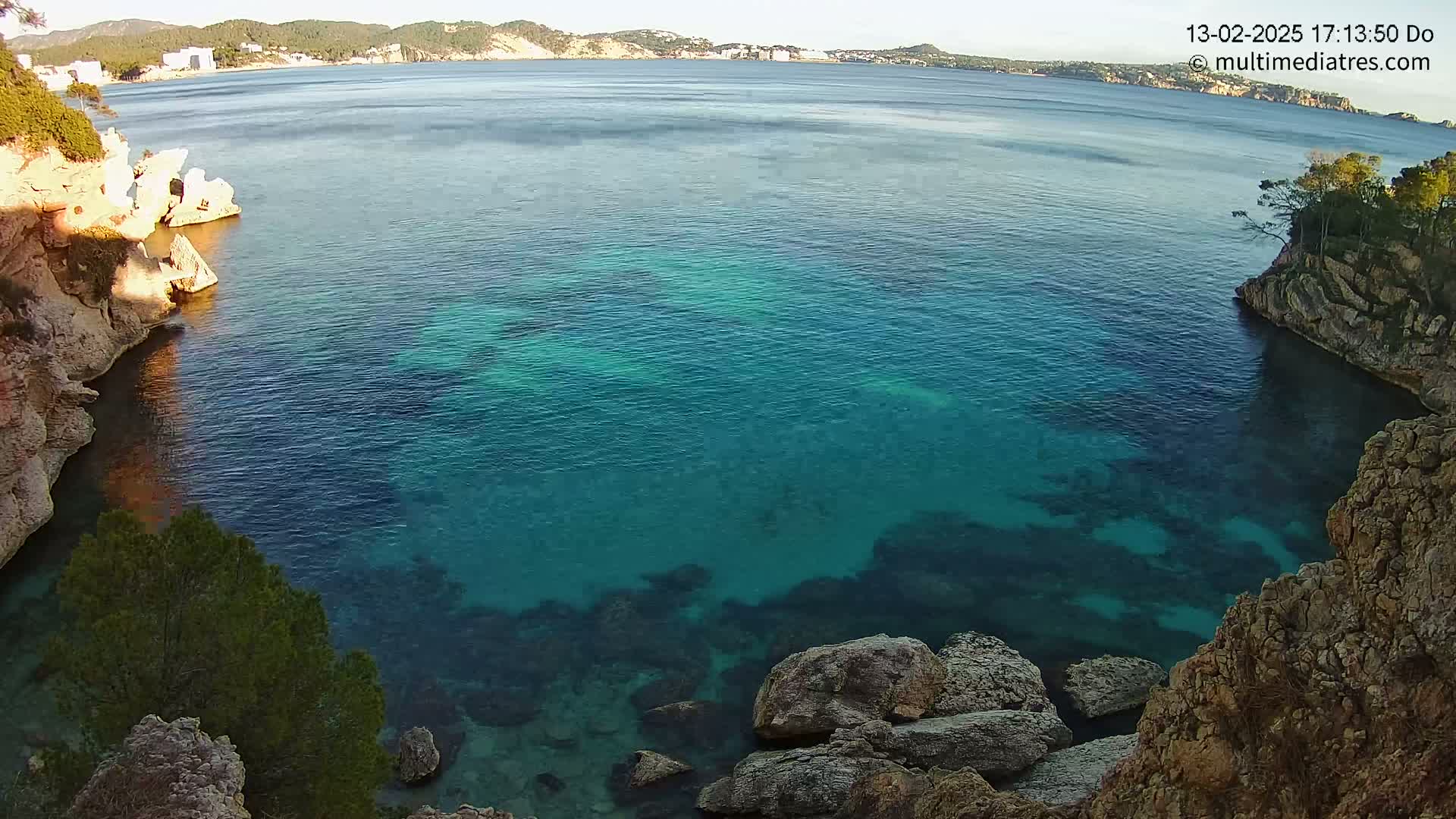 A tranquil cove of clear, teal water is nestled between rocky, tan cliffs on a sunny day, with a distant coastline visible across the bay.