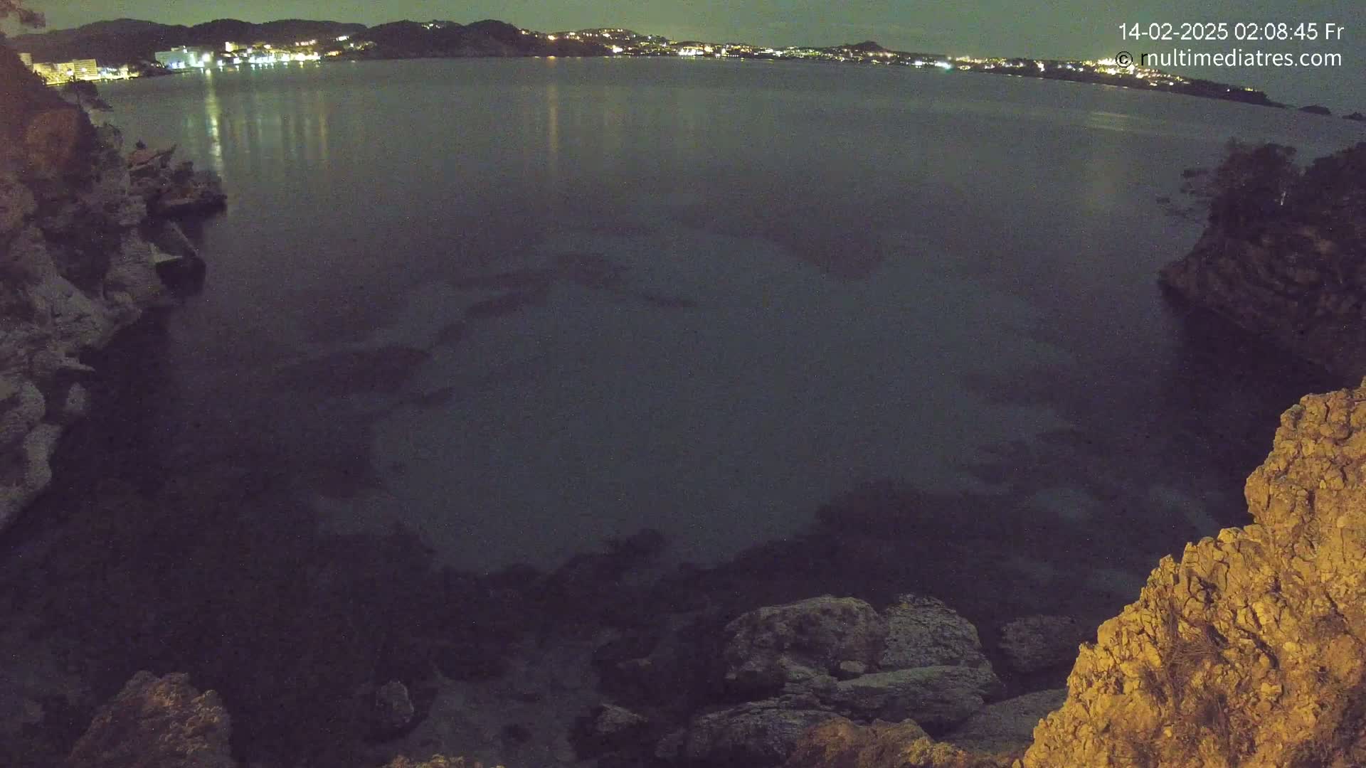 A nighttime view of a dark body of water, partially enclosed by rocky shores, with a town's lights visible in the distance.