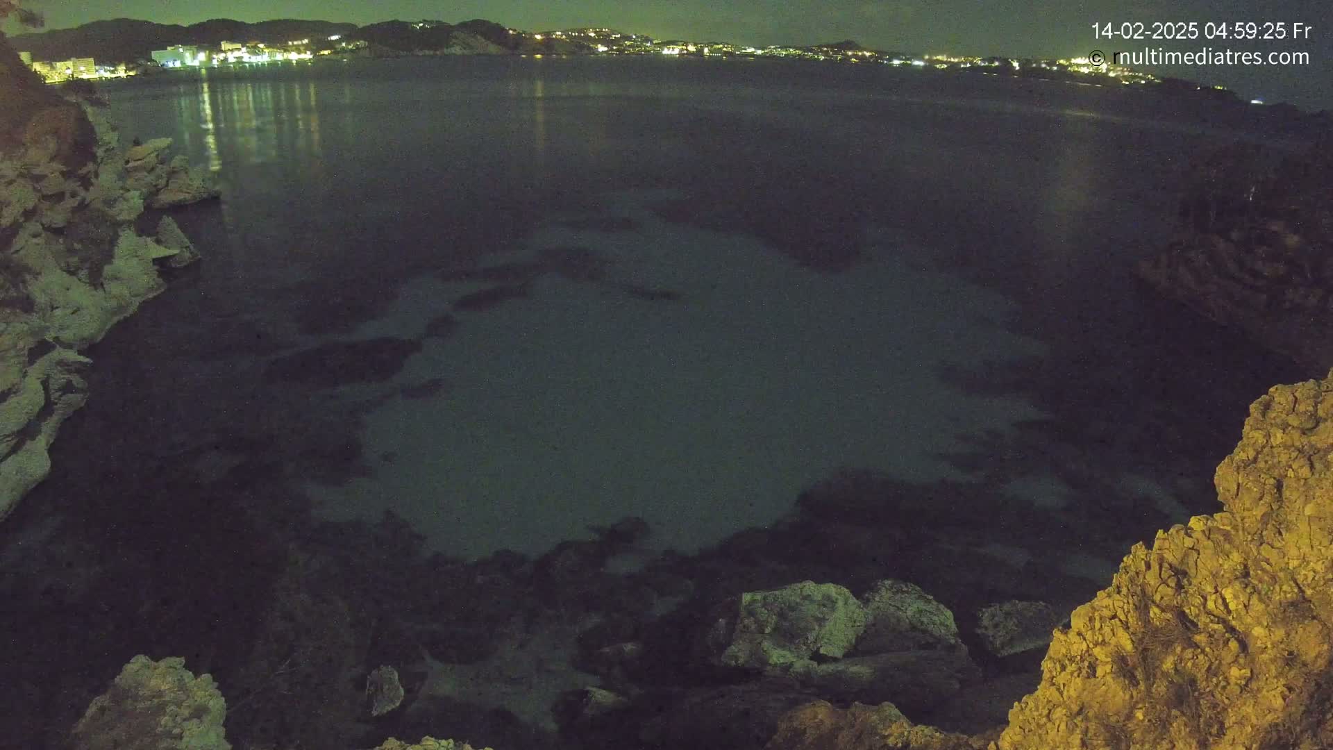 A nighttime view of a dark, calm body of water surrounded by rocky cliffs, with a distant town illuminated by lights across the water.