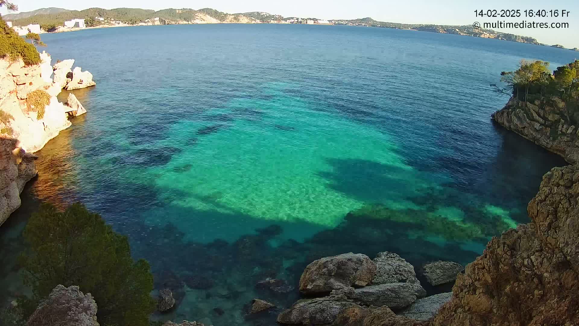 A cove of clear, teal water is nestled between rocky, light-brown cliffs under a bright, sunny sky.