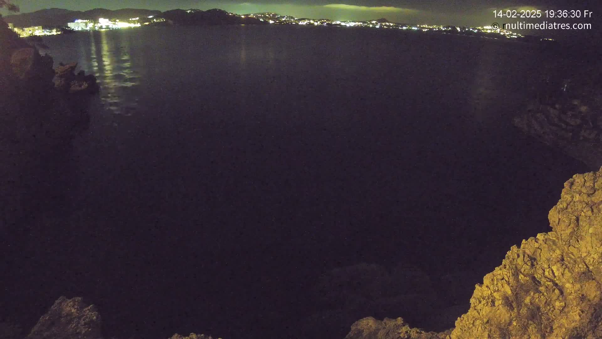 A nighttime view of a dark body of water reflecting city lights in the distance, framed by rocky, land formations.