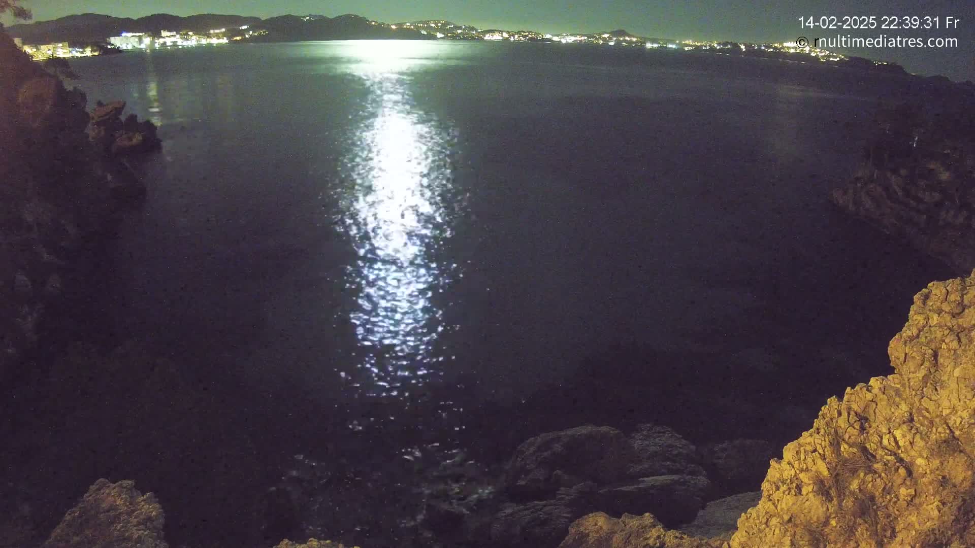 A moonlit night scene shows a calm body of water reflecting light, with dark rocky shoreline in the foreground and a distant city skyline illuminated at night.