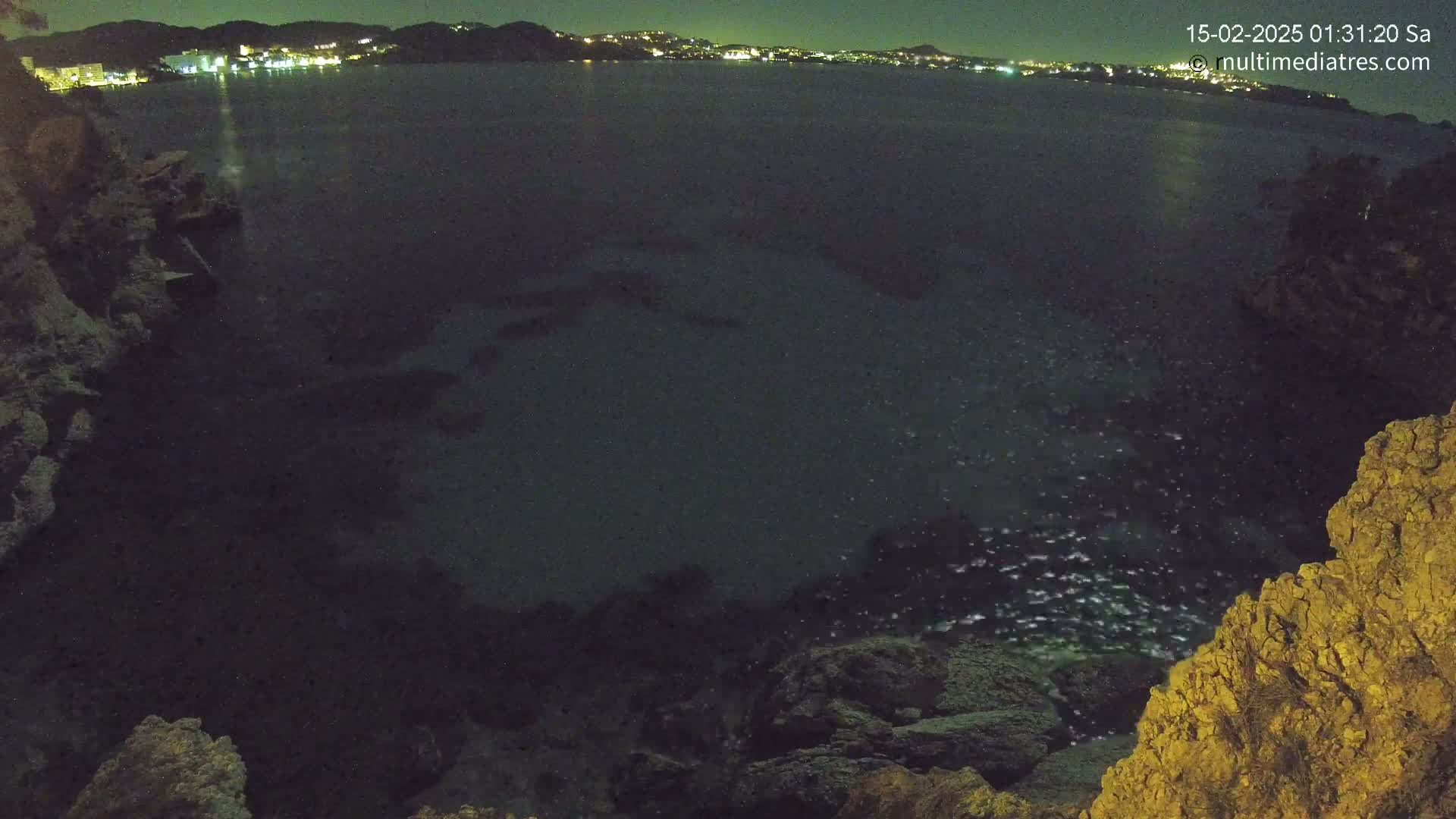 A nighttime view of dark water, illuminated by distant city lights, next to rocky terrain.