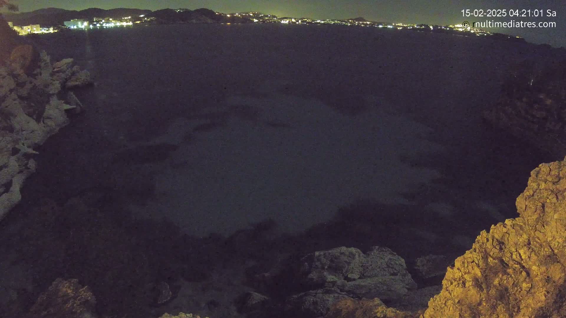 A nighttime view of a dark body of water, with a distant shoreline showing faint lights, seen from a rocky overlook.