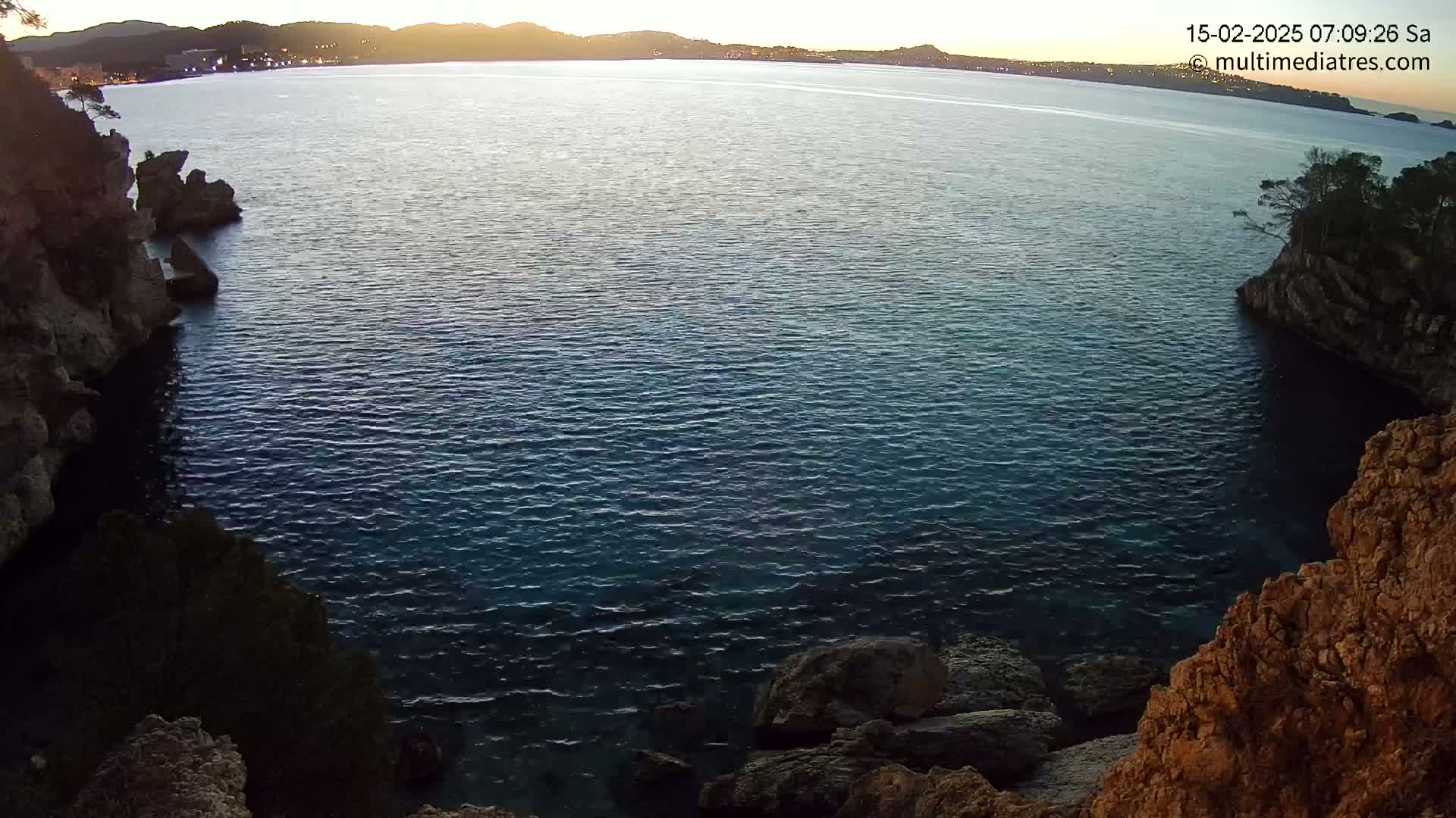 Calm, dark-blue water is nestled between rocky cliffs at sunrise, with a distant shoreline visible in the background.