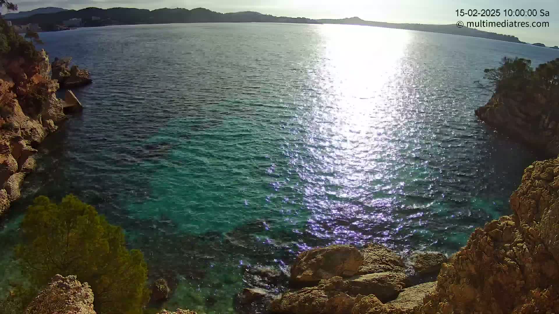 A rocky cove with turquoise water, partially sunlit, overlooks a calm sea and distant hills under a sunny sky.