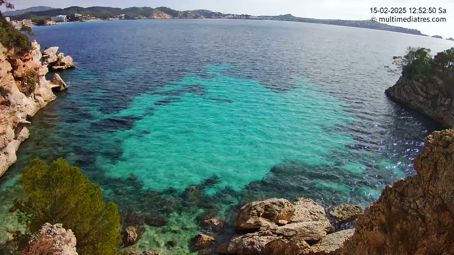 A sunny day reveals a cove of clear, turquoise water nestled between rocky, tan-colored cliffs, with a distant town visible across the bay.