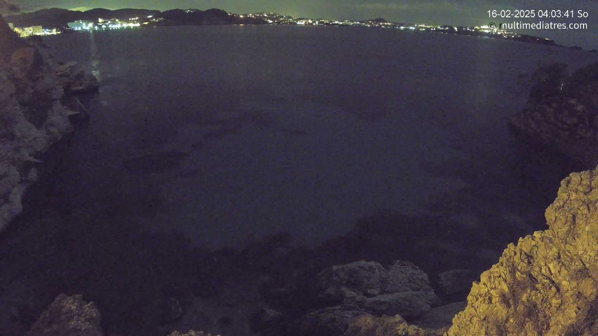 A nighttime view of dark water, illuminated by distant city lights, from a rocky overlook.