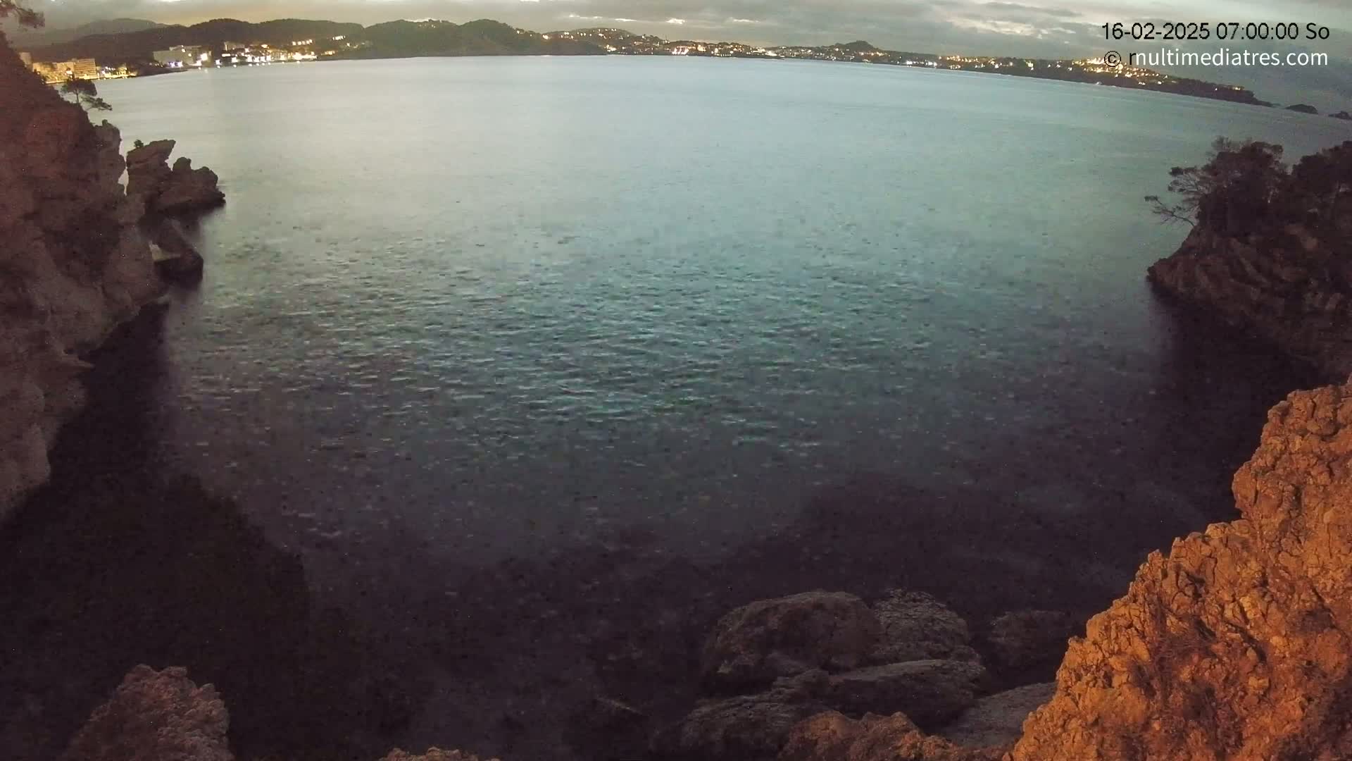 A dark, calm body of water is nestled between rocky cliffs at twilight, with a city's lights visible in the distance.