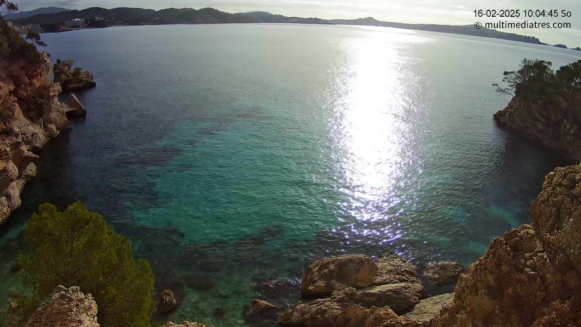 A cove of clear, teal water is surrounded by rocky cliffs and sparse vegetation, with sunlight sparkling on the water's surface under a partly cloudy sky.