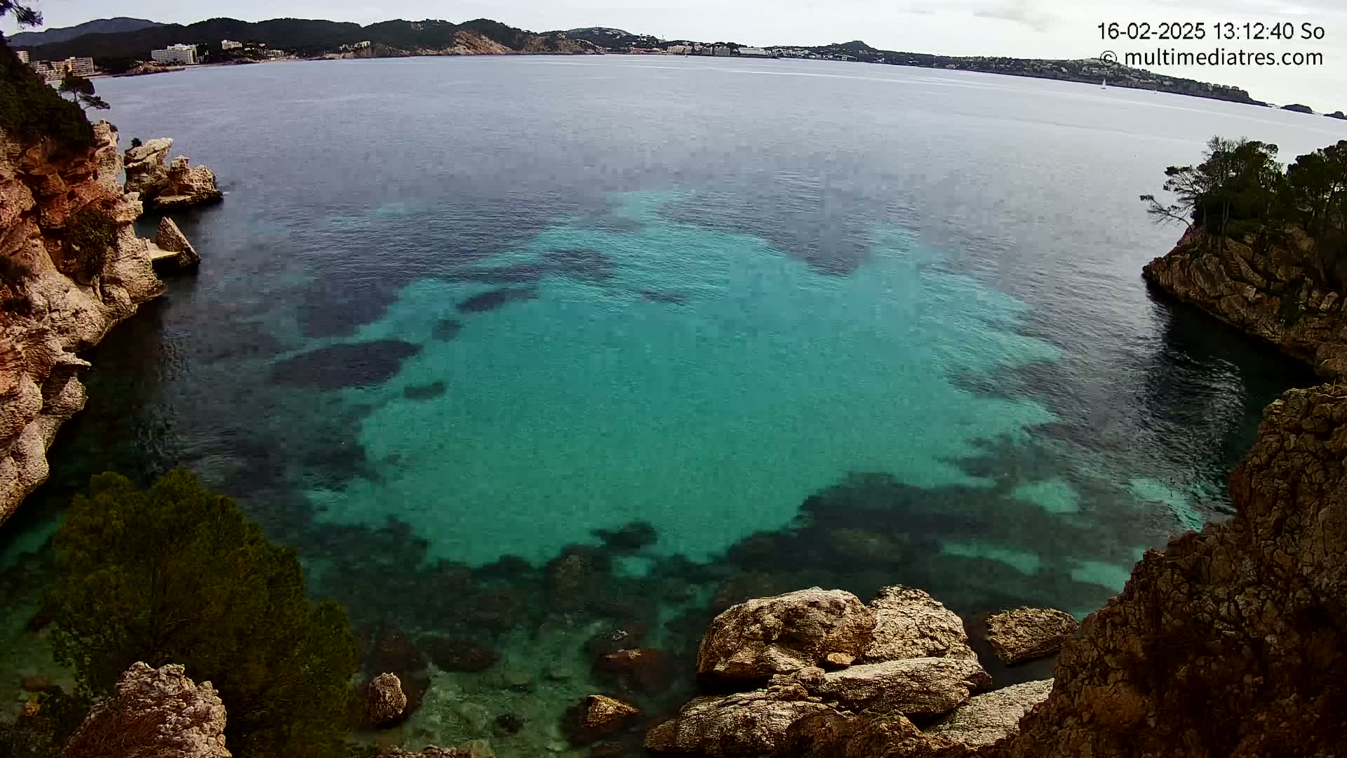 A cove of clear, teal water is nestled between rocky, tan cliffs under a partly cloudy sky.