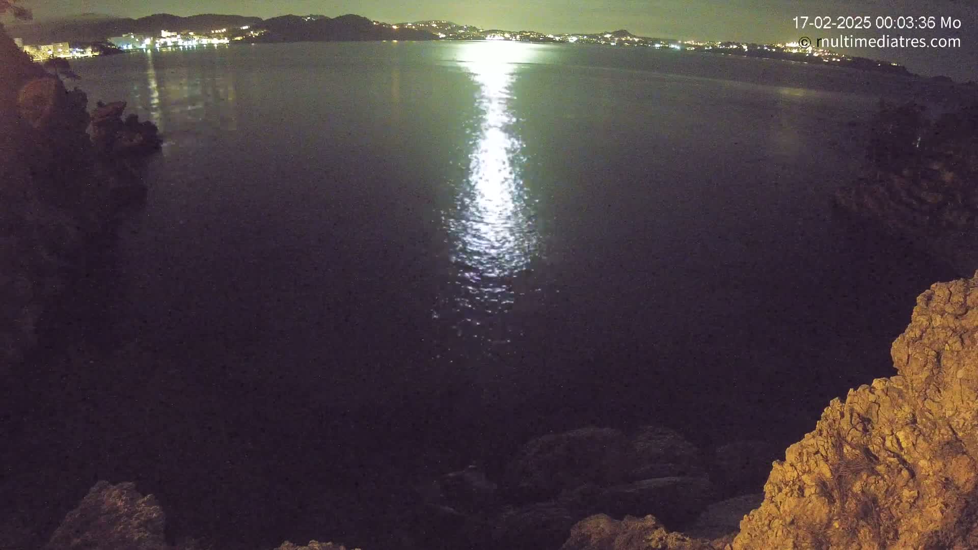 A moonlit night scene shows a dark body of water reflecting light, with a distant, illuminated shoreline visible in the background, and rocky outcrops in the foreground.