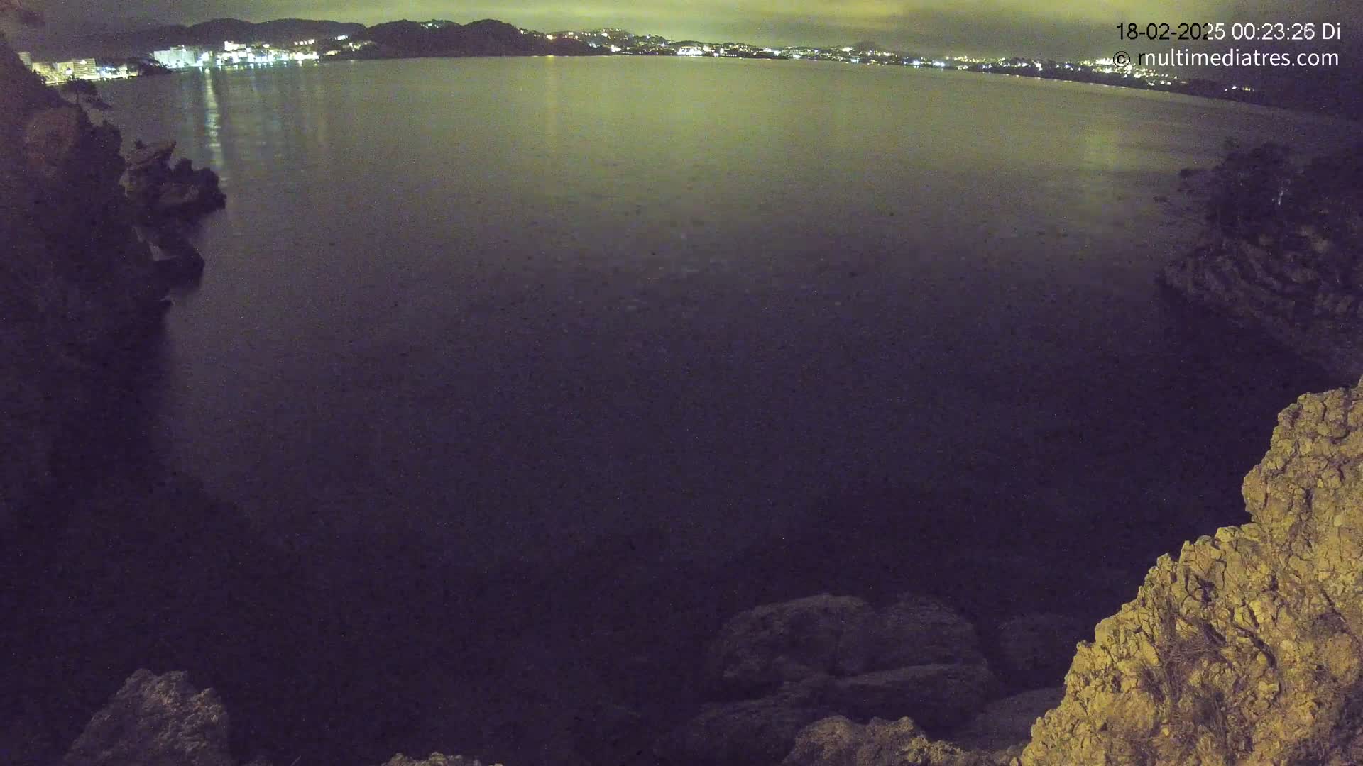 A nighttime view of a dark body of water, illuminated by distant city lights, is seen from a rocky, high vantage point.