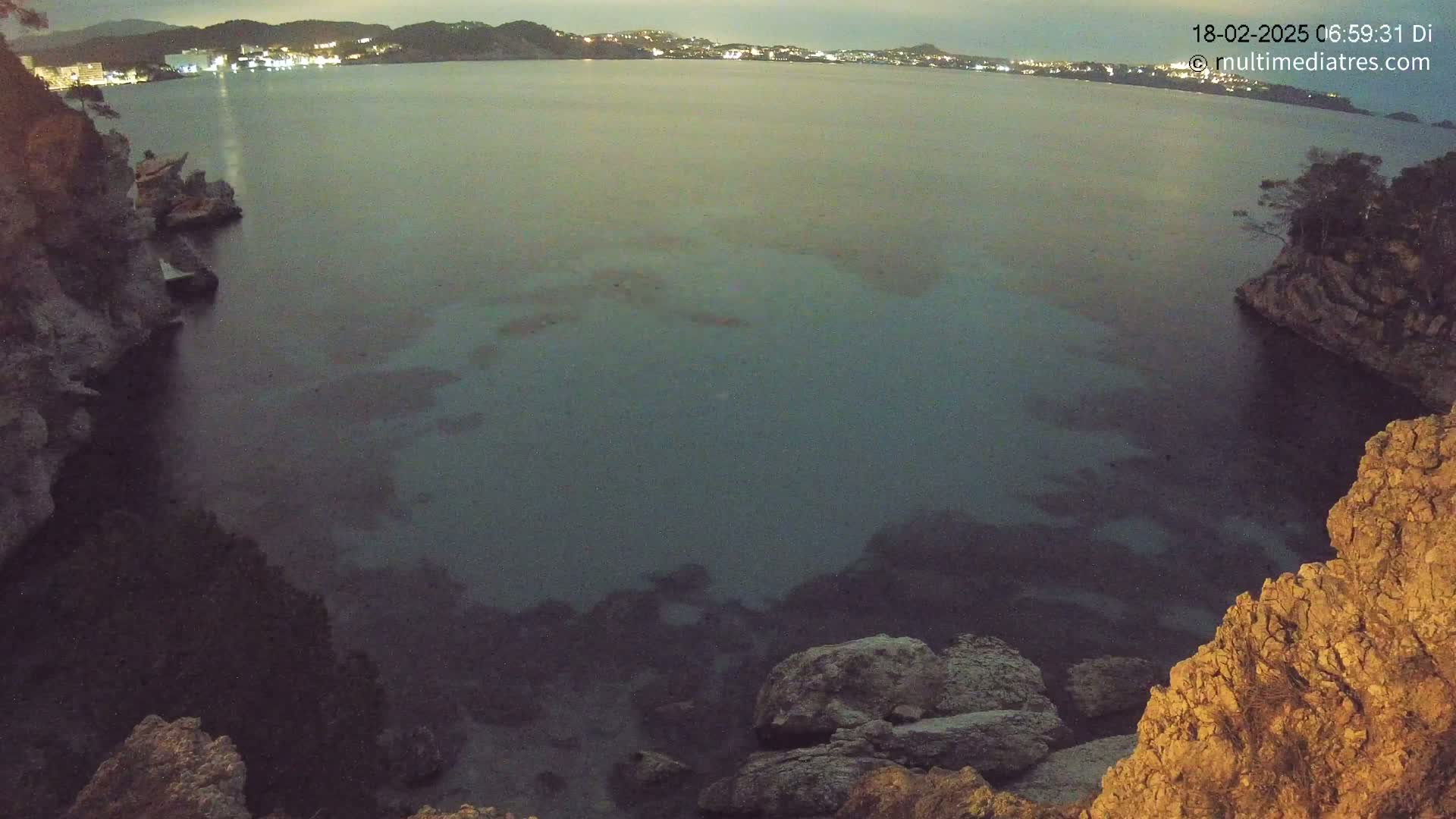 A dark, calm body of water at night is nestled between rocky cliffs, with a distant town faintly illuminated in the background.