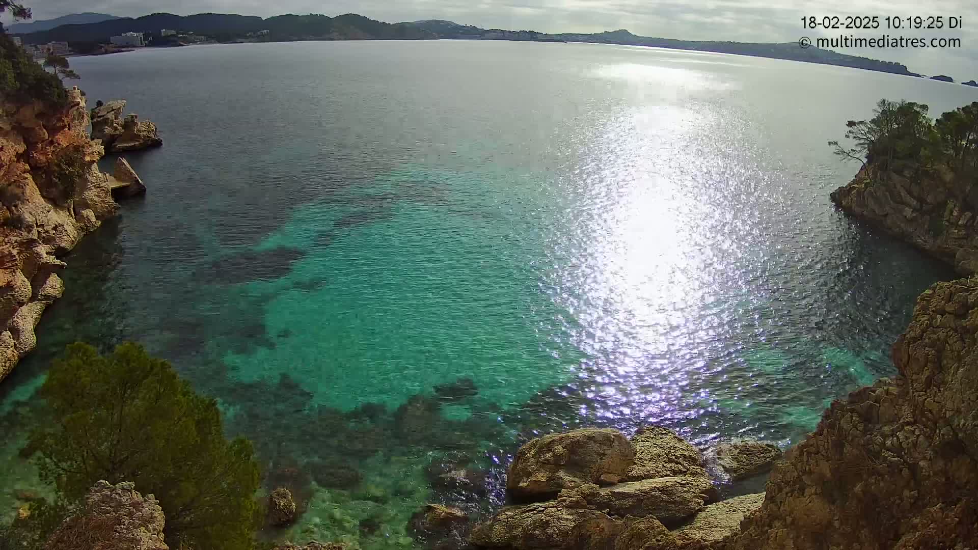 Clear, bright, sunny weather illuminates a cove of sparkling turquoise water surrounded by rocky, tan cliffs and evergreen trees.