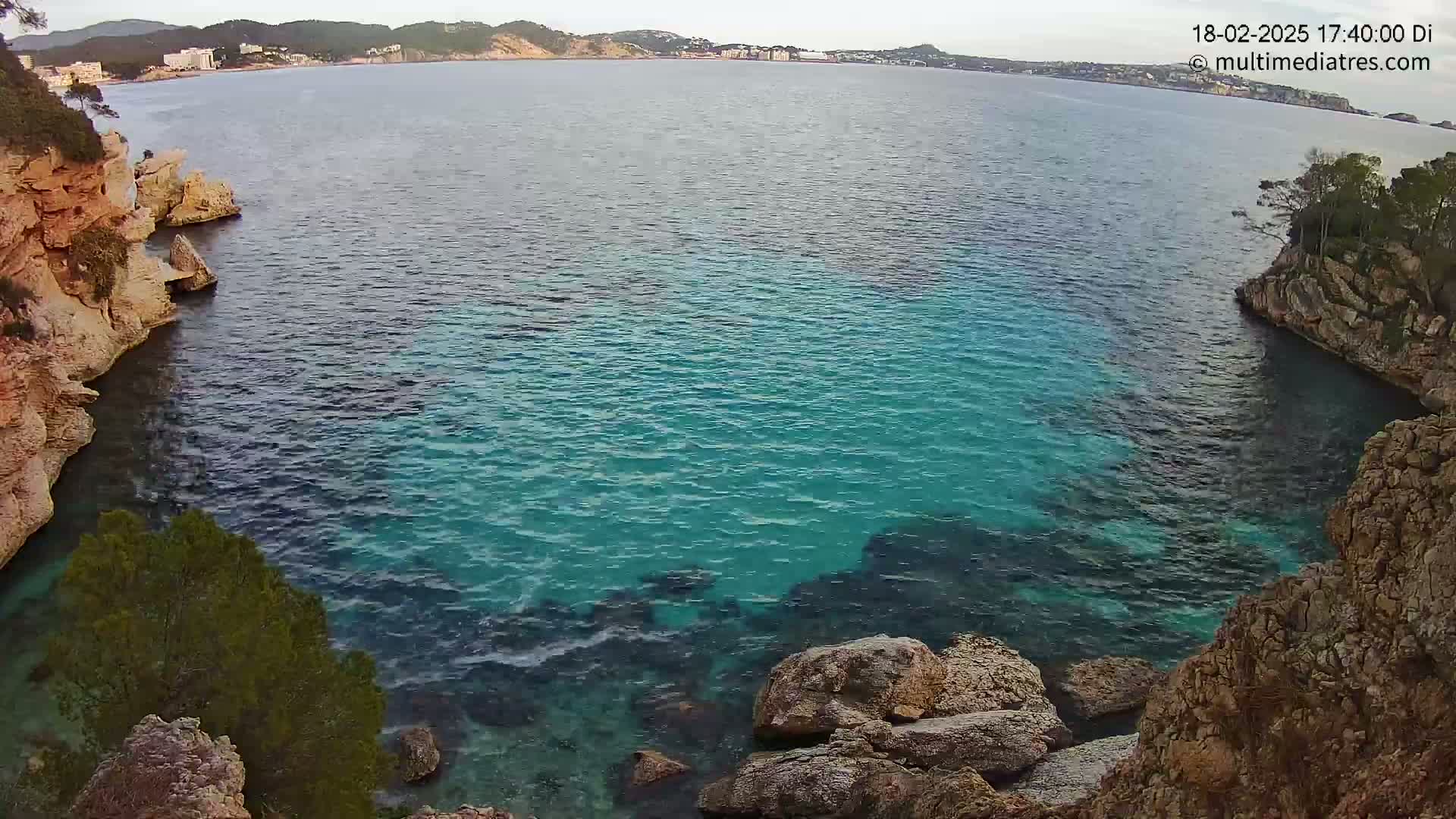 Clear, teal water fills a cove between rocky, lightly vegetated cliffs, with a calm sea and distant coastline visible in the background.