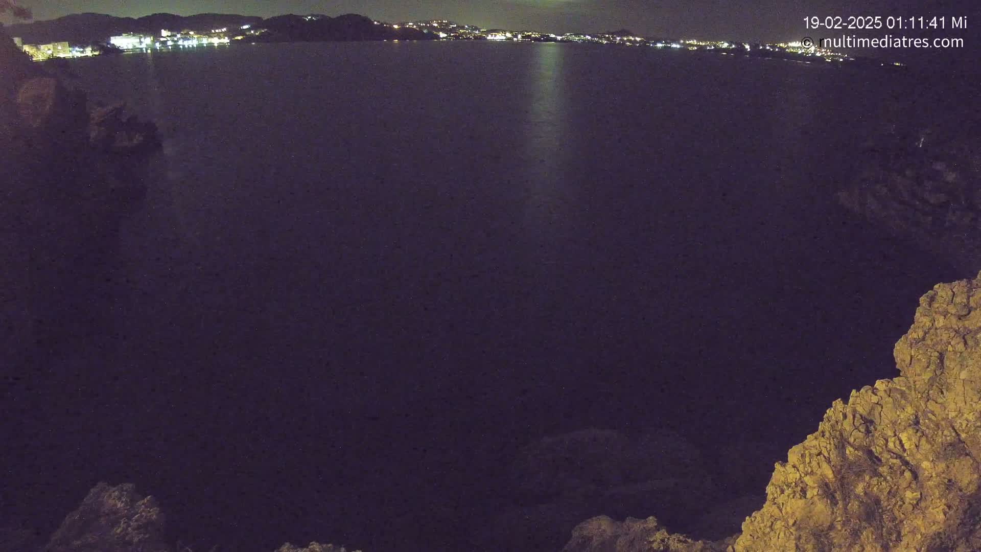 A nighttime view of a dark body of water reflecting city lights on the horizon, framed by rocky, land formations.