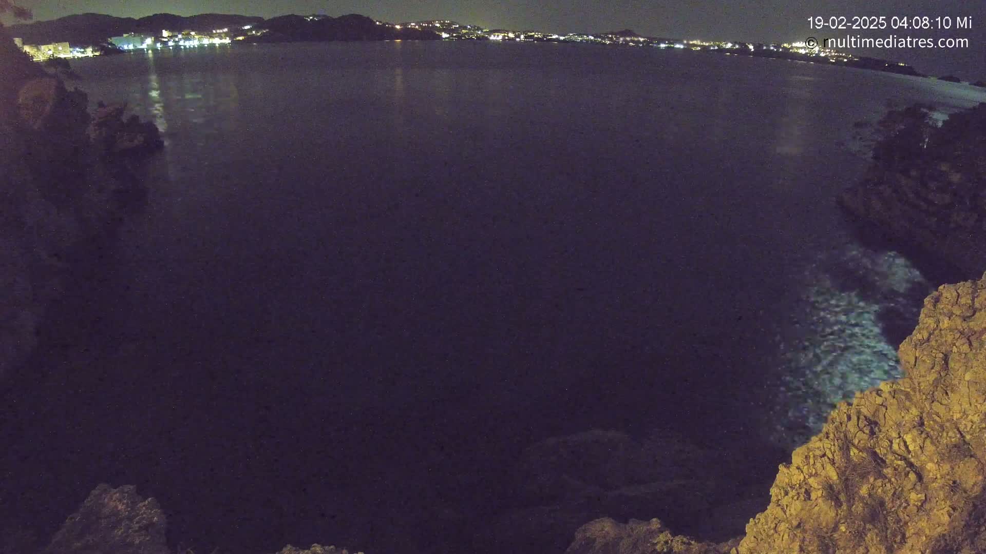 A nighttime view of a dark body of water reflecting the lights of a distant town, framed by rocky terrain.