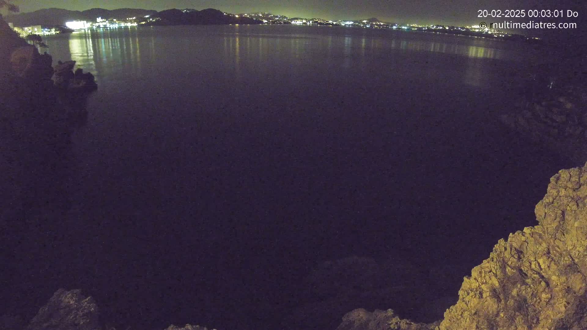 A nighttime view of a calm body of water reflecting the lights of a city in the distance, seen from a rocky vantage point.