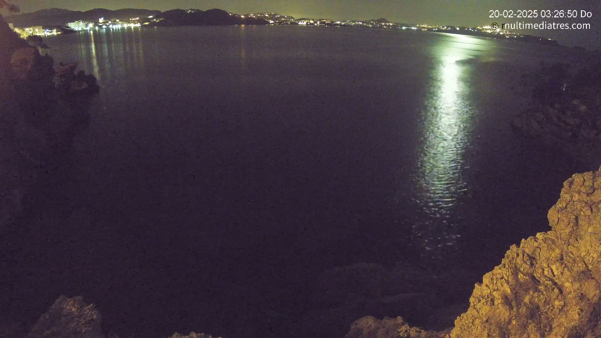 A nighttime view of a calm body of water reflecting the lights of a distant town, seen from a rocky vantage point.