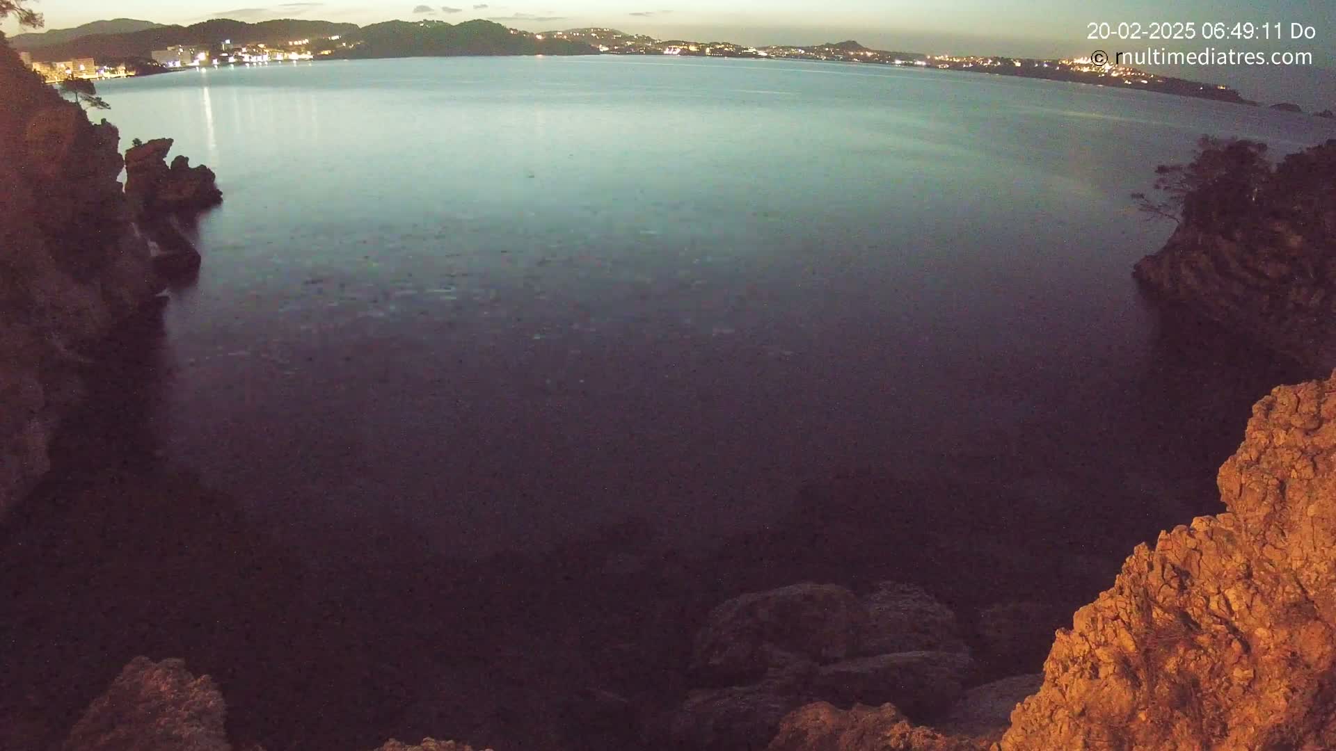 A dark, calm body of water is seen at twilight, bordered by rocky, craggy shorelines, with a town faintly lit in the distance.