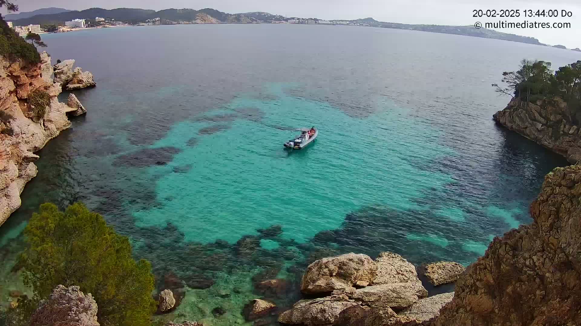 A small motorboat sits in a cove of clear, turquoise water surrounded by rocky, tan-colored cliffs and greenery under a partly cloudy sky.