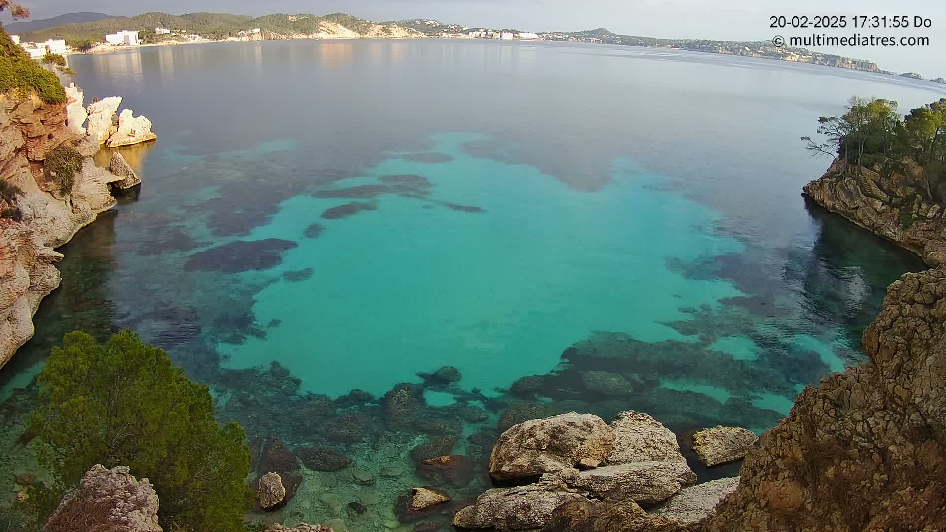 A cove of clear, turquoise water is nestled between rocky, vegetated cliffs, with a calm sea and distant shoreline visible in the background.