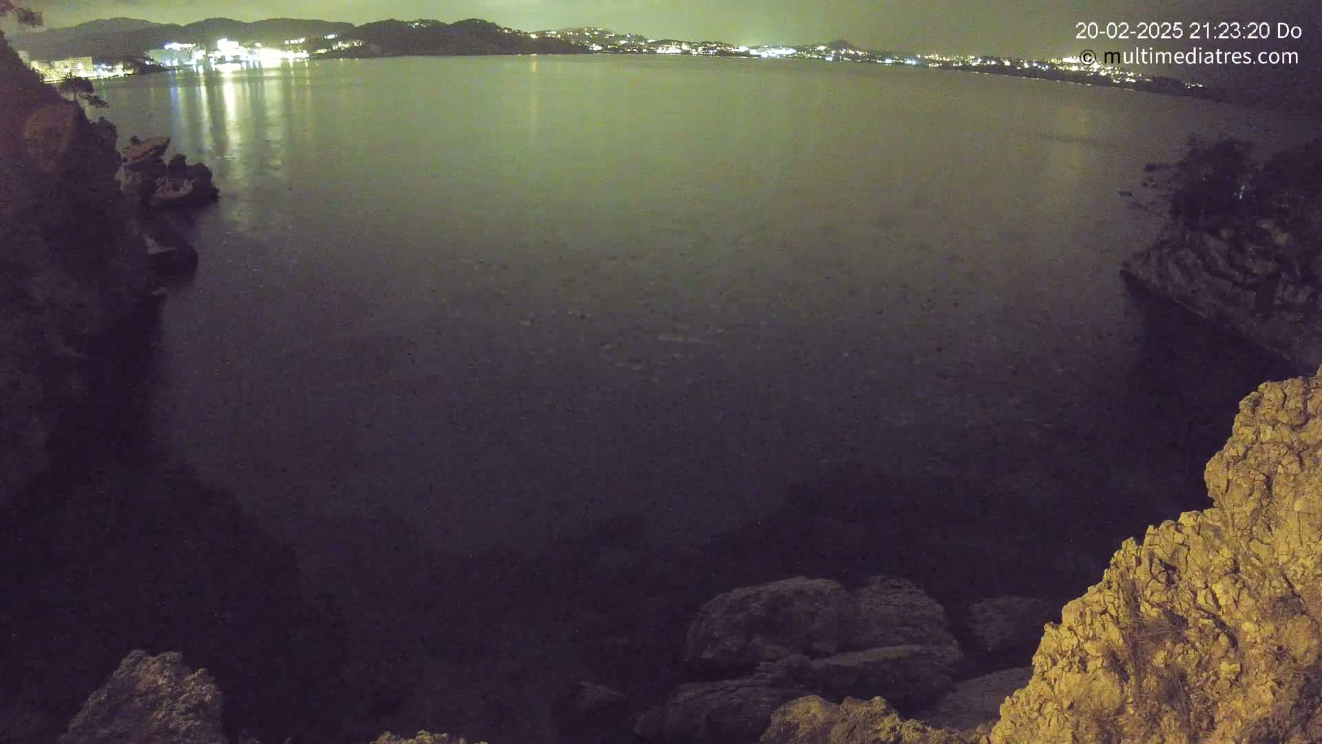 A nighttime view of a dark body of water, reflecting the lights of a distant town across the water, is seen from a rocky cliff.