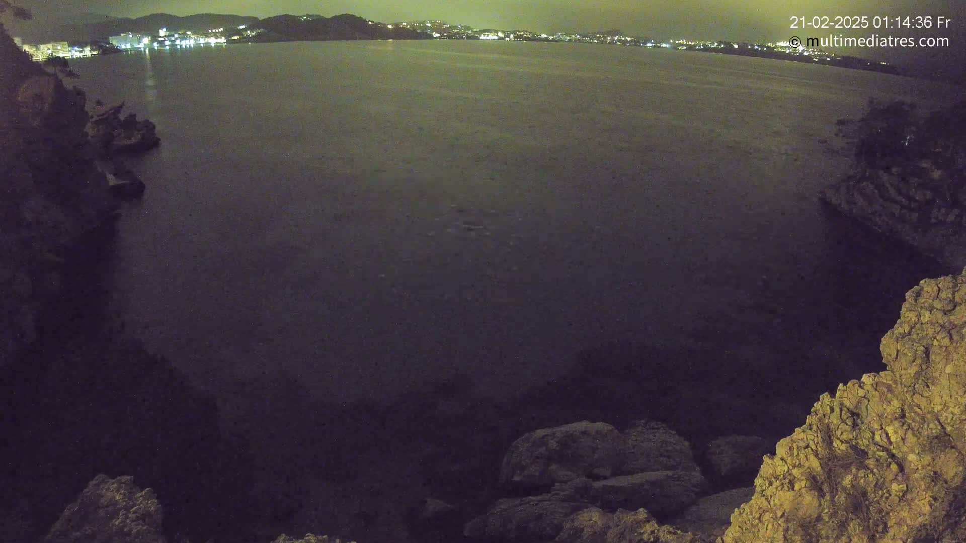 A nighttime view of a dark body of water with a city's lights visible in the distance, framed by rocky cliffs.