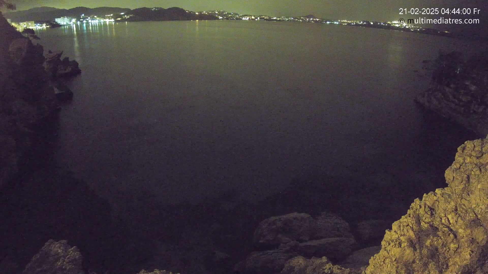 A dark, nighttime view of a calm body of water with a distant, illuminated town shoreline visible from a rocky outcrop.