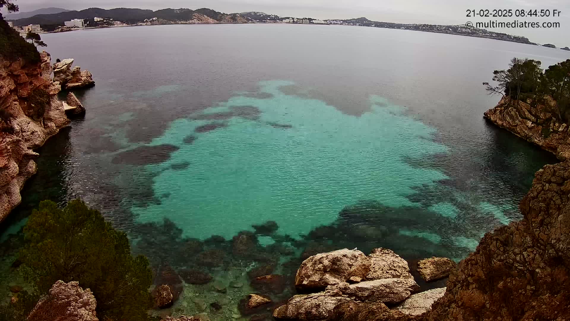 A cove of teal-colored water is nestled between rocky cliffs on an overcast day, with a distant town visible across the bay.