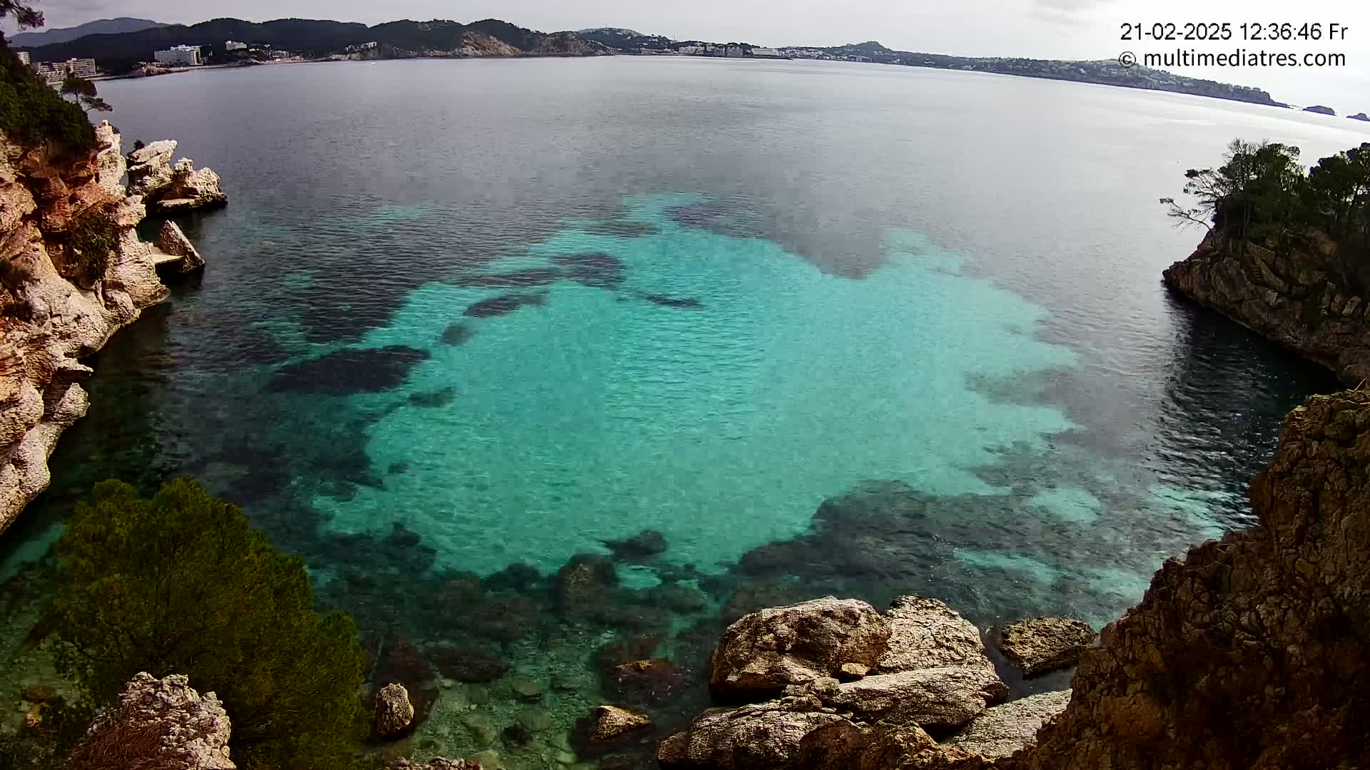 A cove with clear, turquoise water is nestled between rocky cliffs on a partly cloudy day.
