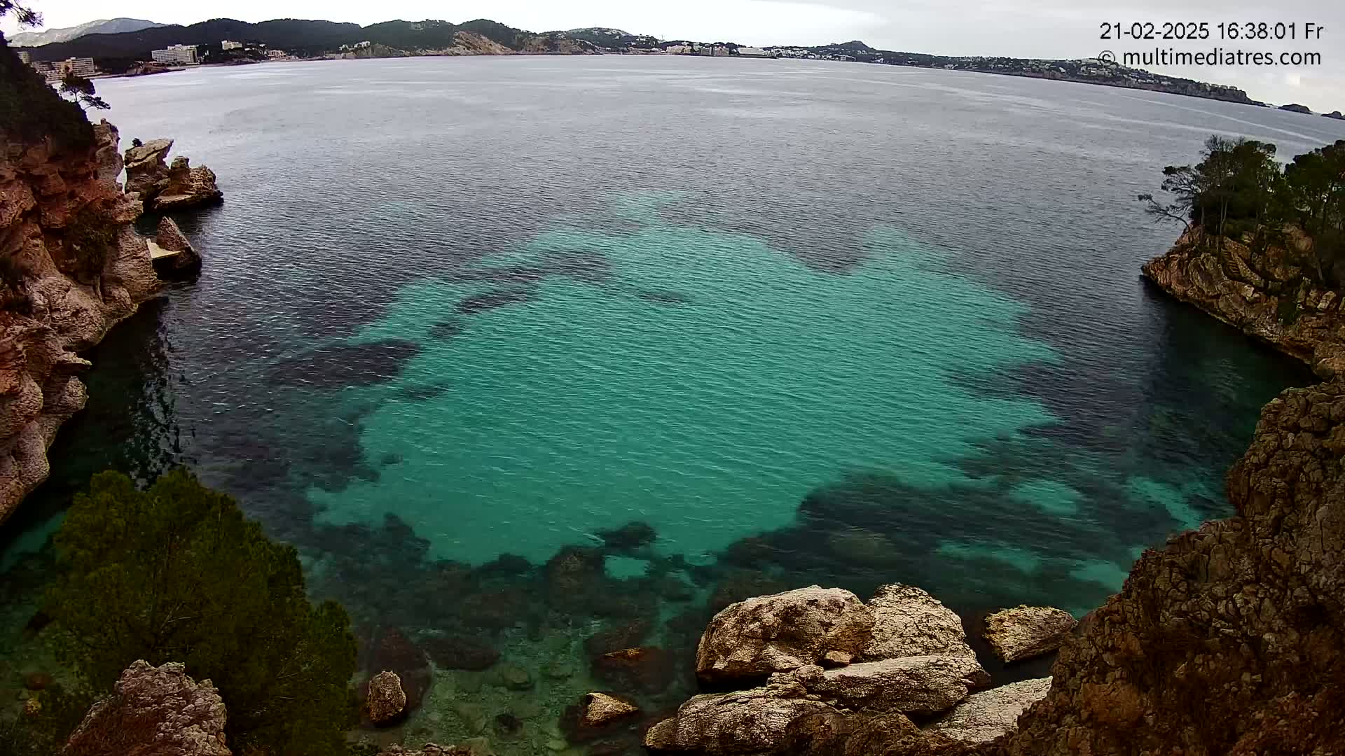 A cove of teal-colored water is nestled between rocky, reddish-brown cliffs on an overcast day.