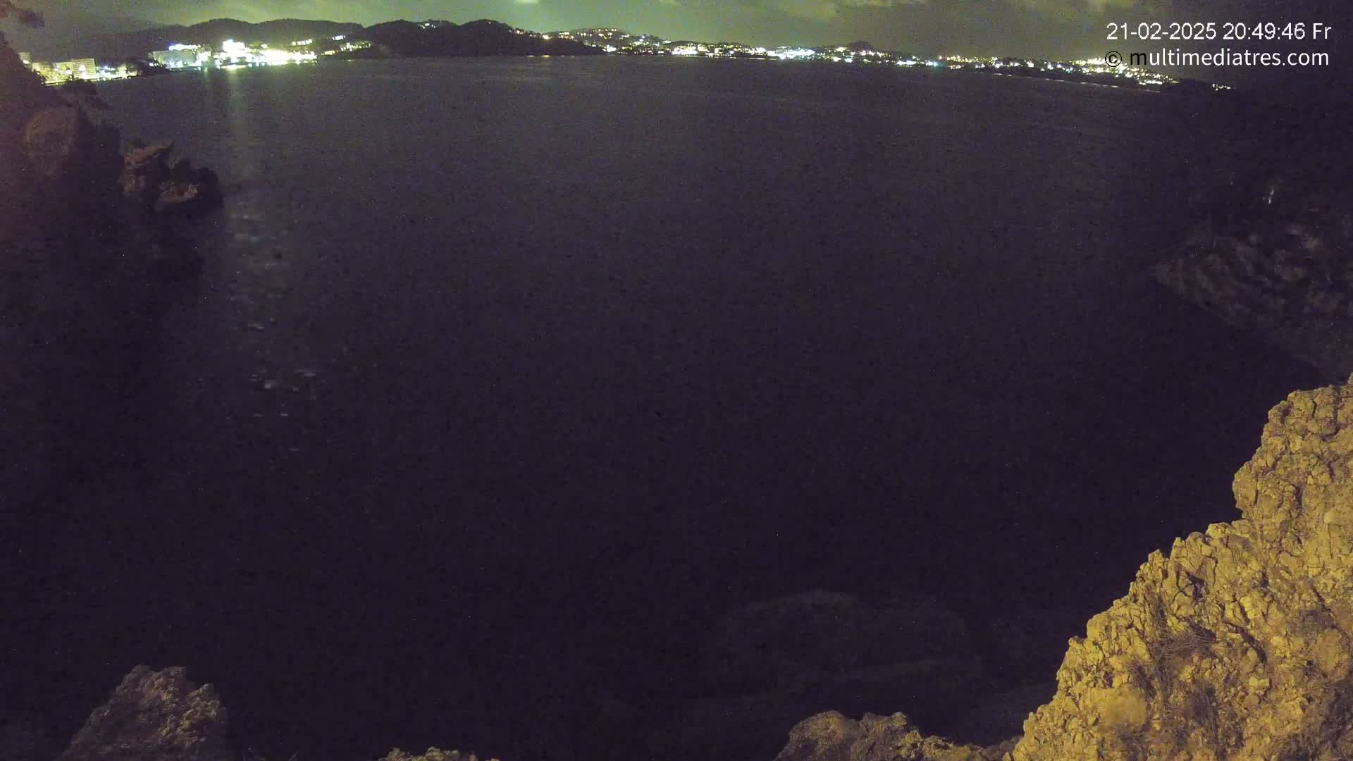 A nighttime view shows a dark body of water with a distant, brightly lit town visible in the background, framed by rocky terrain in the foreground.