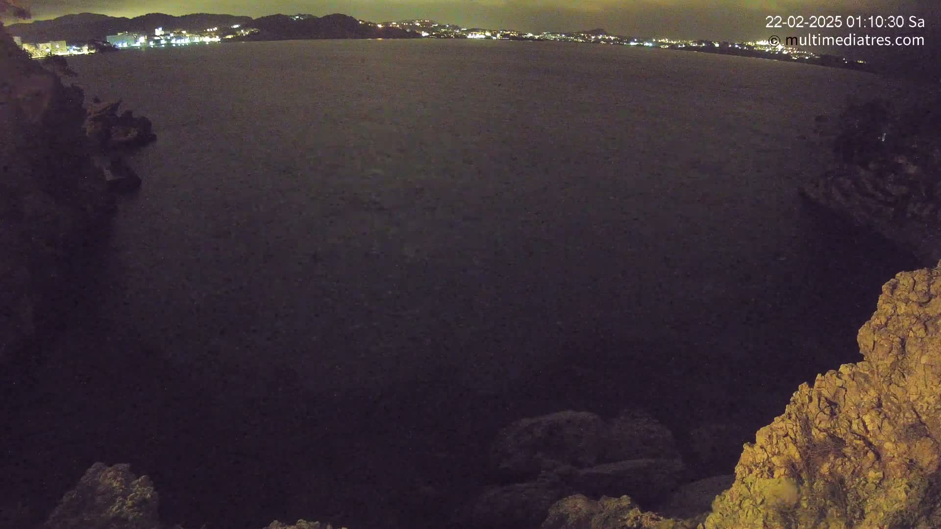 A nighttime view of a dark body of water with a distant, illuminated cityscape and rocky terrain in the foreground.