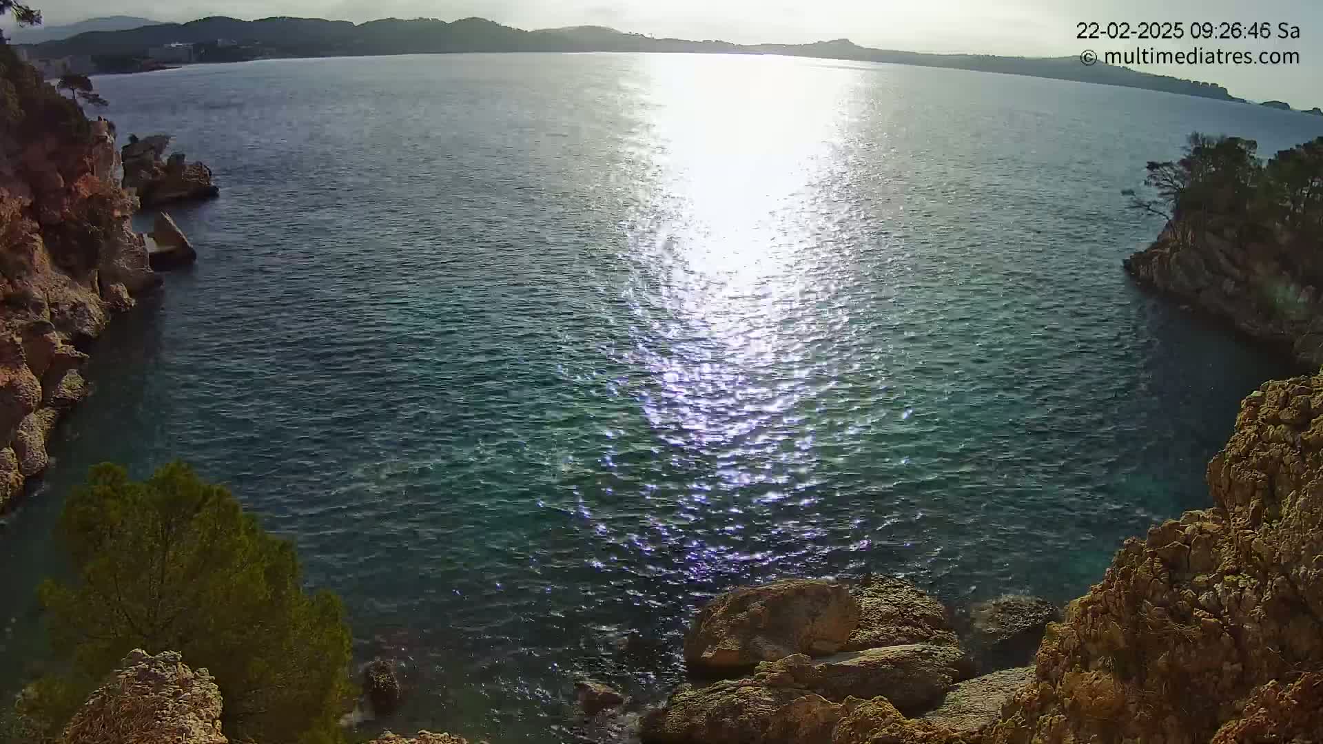 A sunny day reveals a calm, glistening sea nestled between rocky, vegetated cliffs.