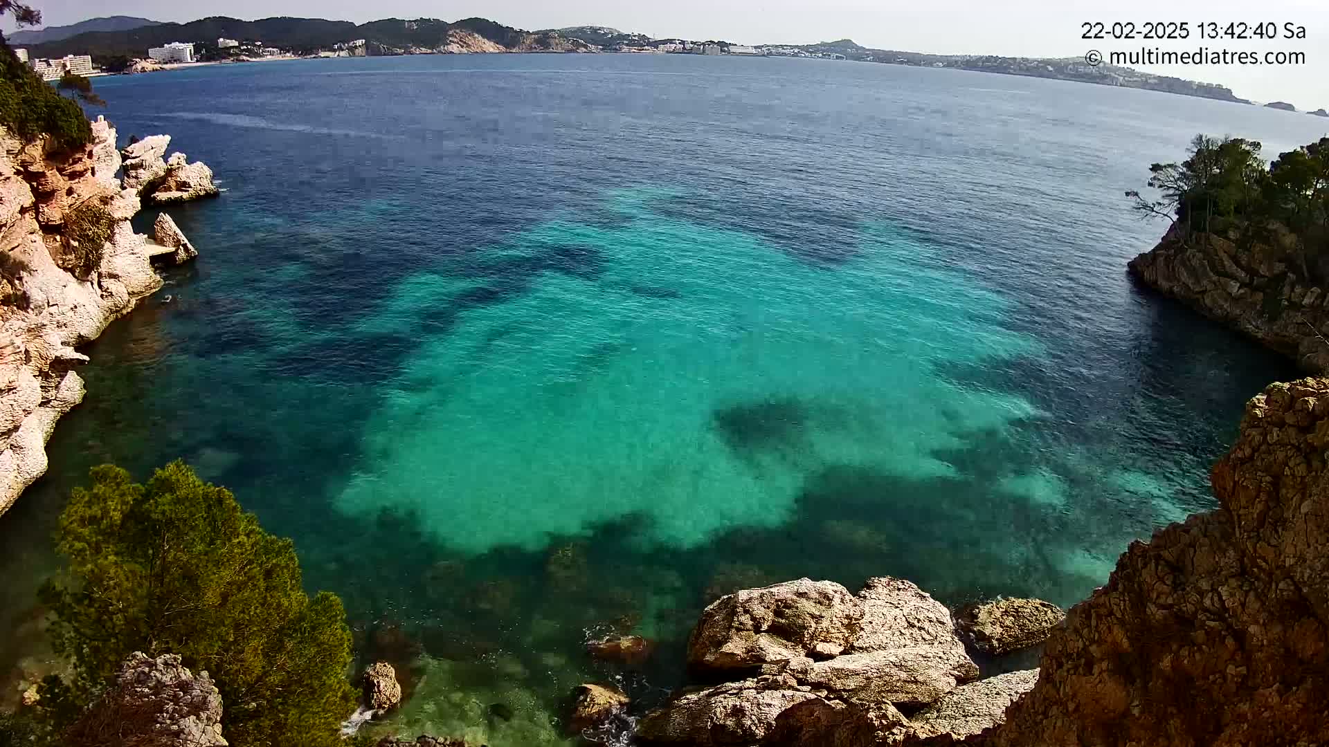 Clear, teal water fills a cove between rocky cliffs on a sunny day, with a distant view of a coastline.
