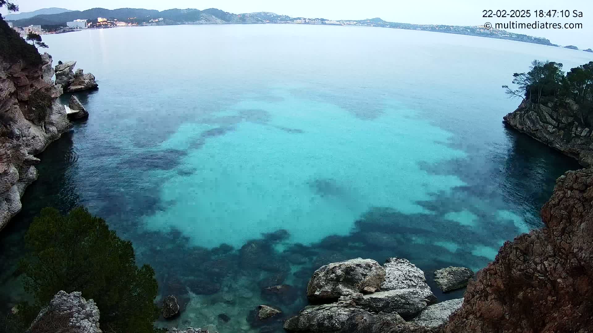 A tranquil cove of clear, teal water is nestled between rocky, tree-covered cliffs under a cloudy sky.