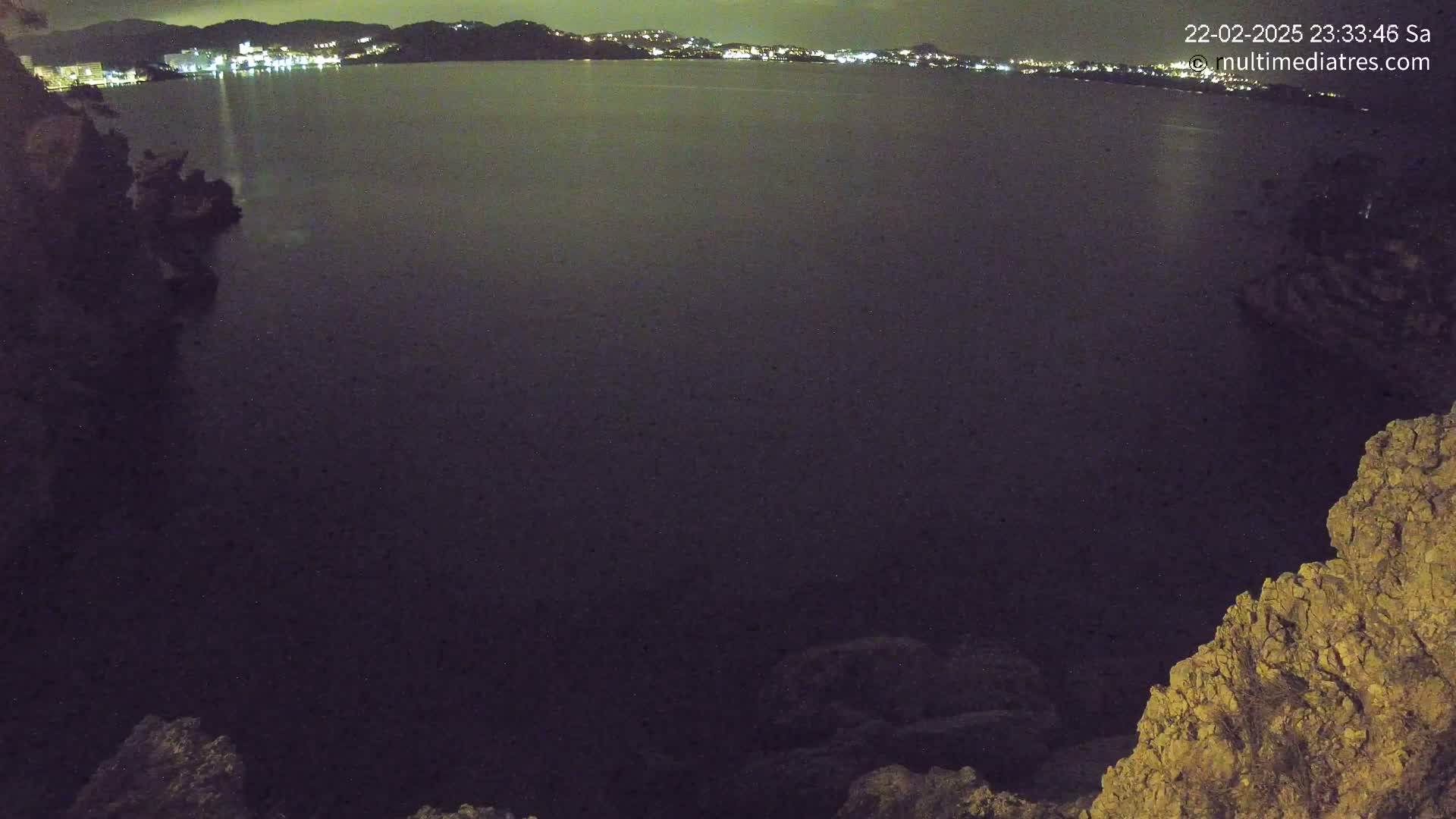 A nighttime view of a dark body of water with a distant, illuminated town visible across the water, framed by rocky terrain.