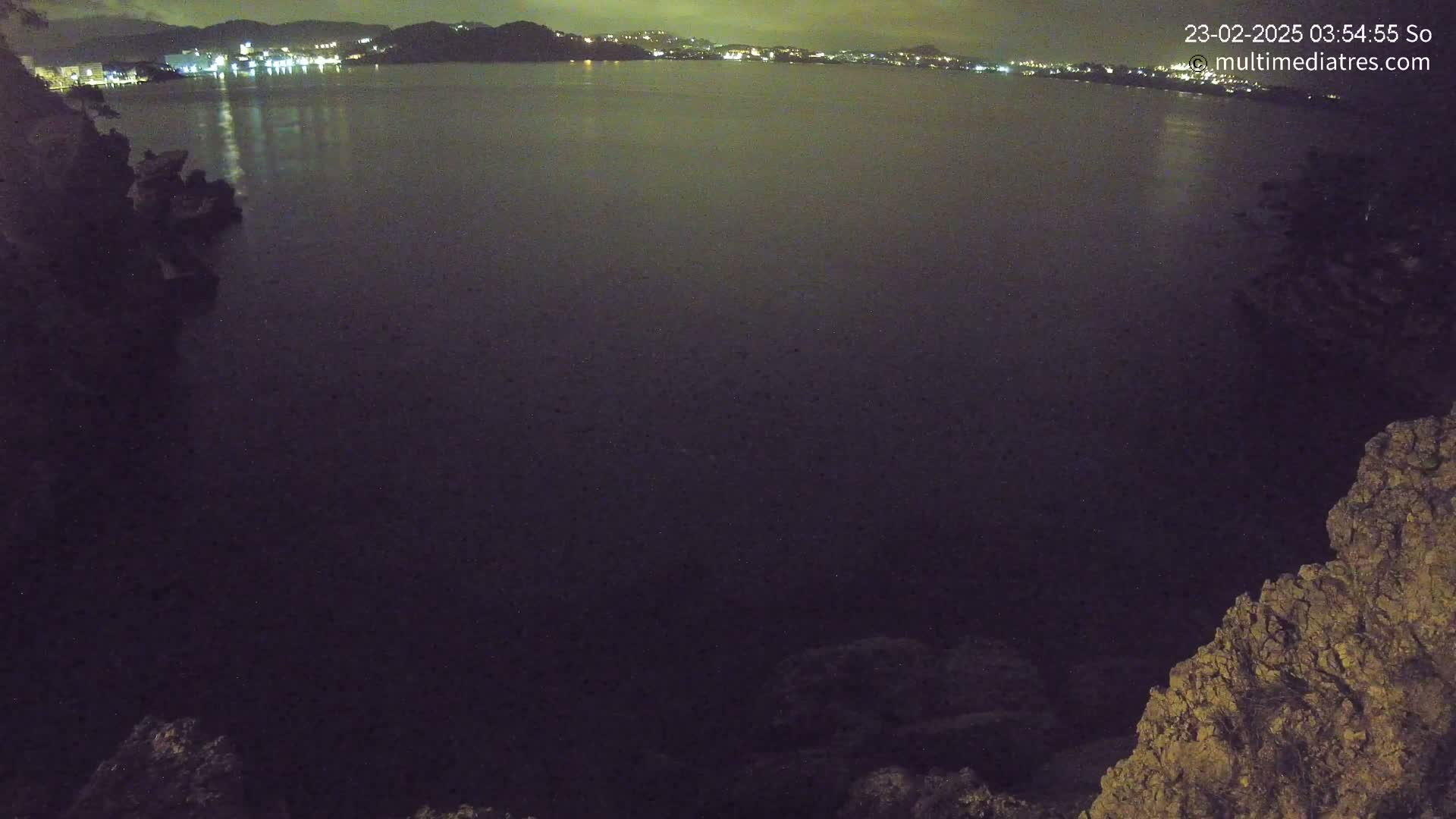 A dark body of water at night is seen from a high vantage point on rocky terrain, with a distant city illuminated on the opposite shore.