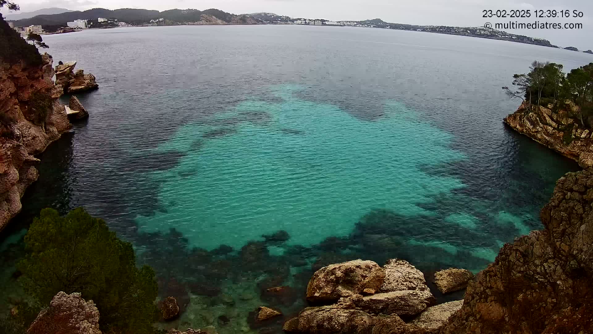 A cove of teal water is nestled between rocky, tan-colored cliffs on an overcast day, with a town visible in the distance across the bay.