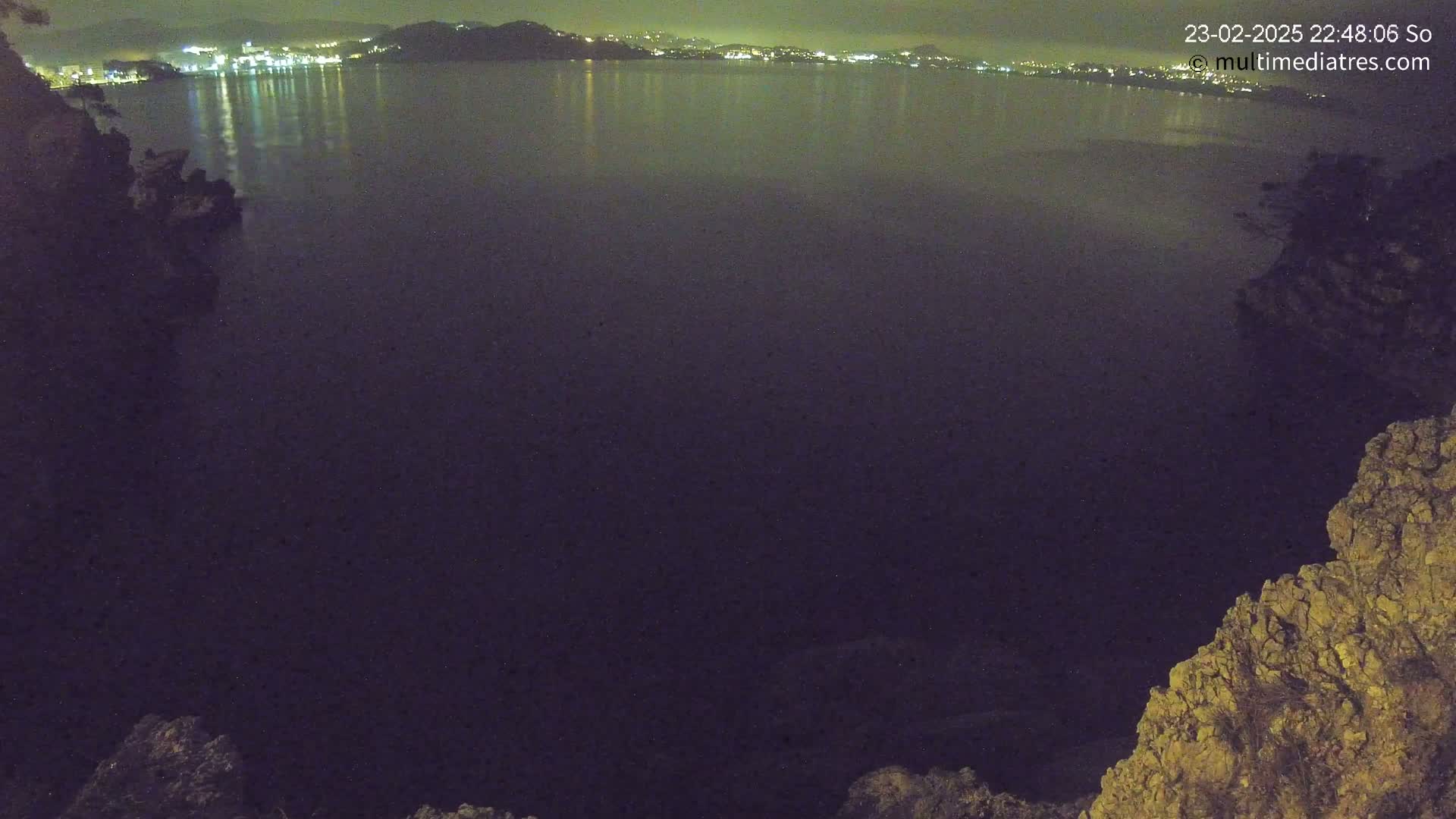 A nighttime view of a calm body of water reflecting the lights of a city on the far shore, seen from a rocky vantage point.
