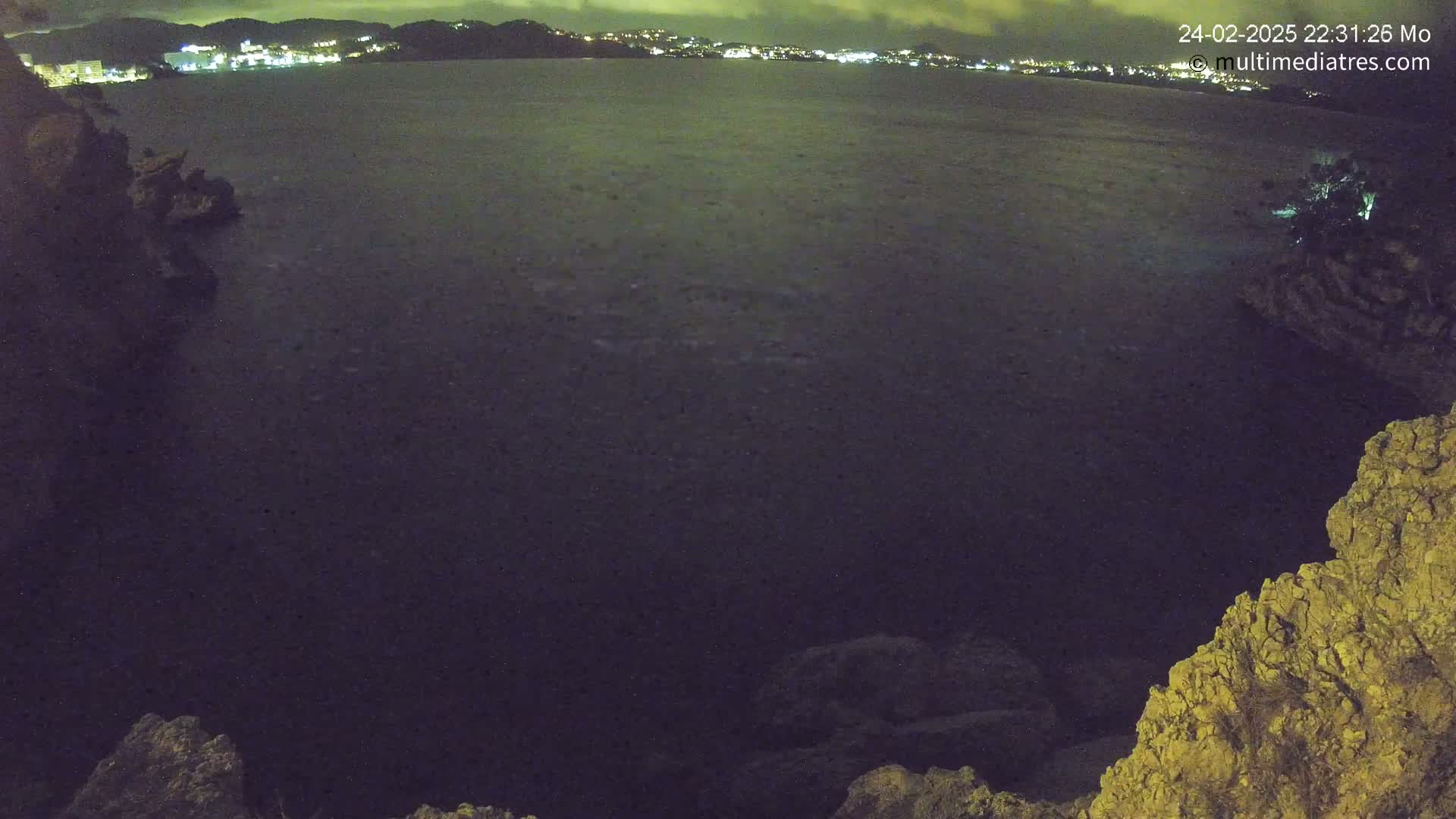 A nighttime view of dark water, partially illuminated by city lights in the distance, from a rocky vantage point.
