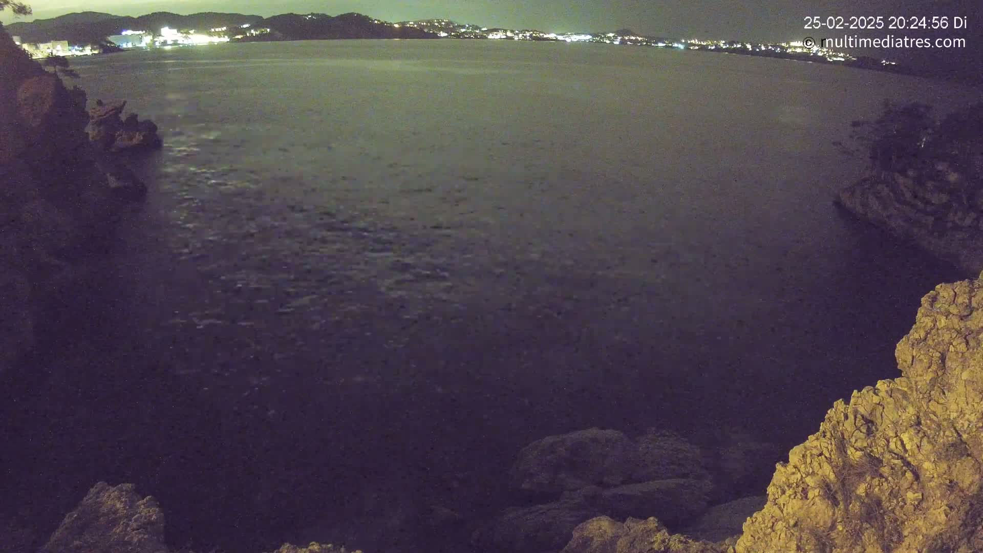 A nighttime view of a dark body of water, possibly the sea, with rocky cliffs on either side and a distant, lit shoreline.