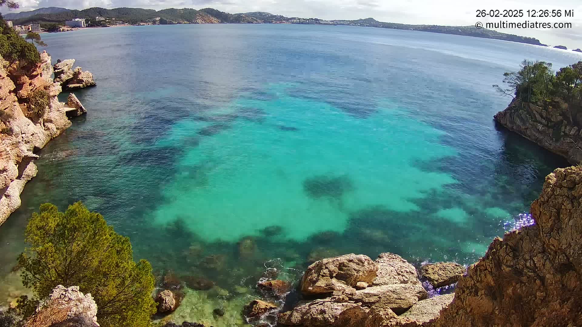 A cove of bright turquoise water is nestled between rocky, tan cliffs under a mostly sunny sky.